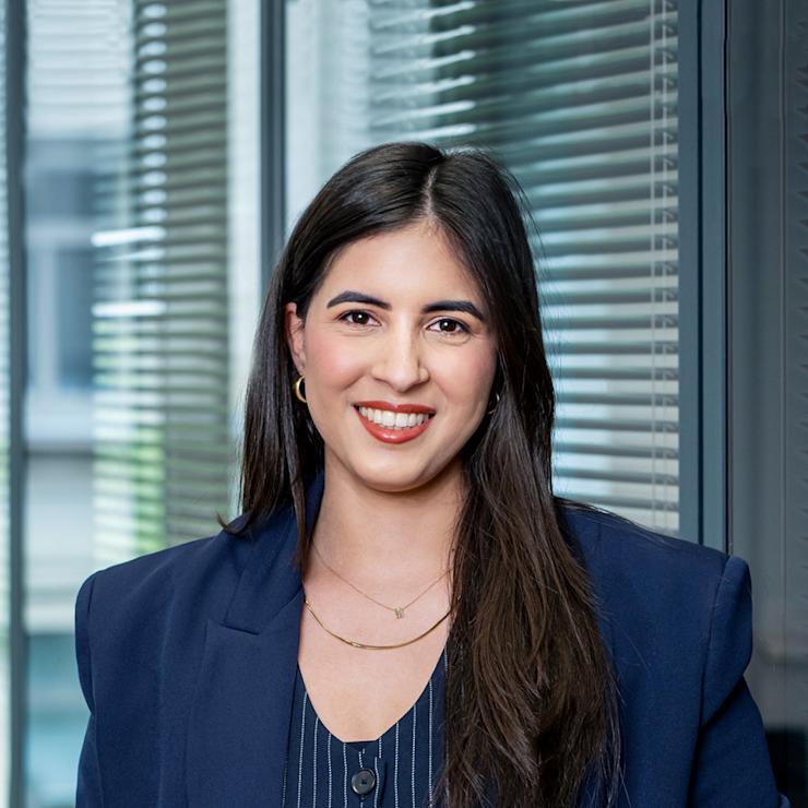 Junge Frau mit dunklen Haaren steht in einem Büro vor Glas und Jalousien.