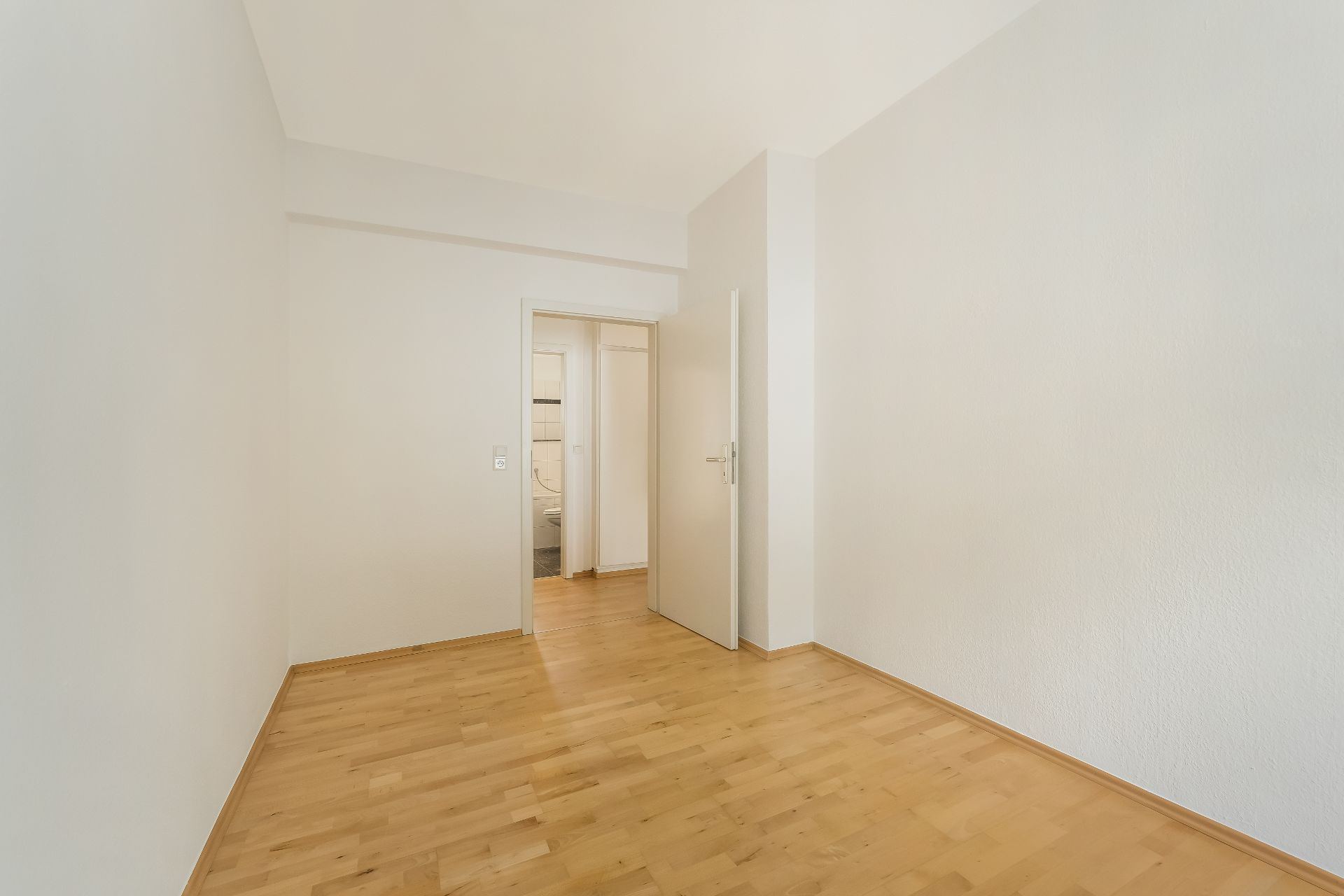 Leerer Raum mit hellem Holzboden und weißen Wänden, geöffnete Tür führt zu weiterem Raum.