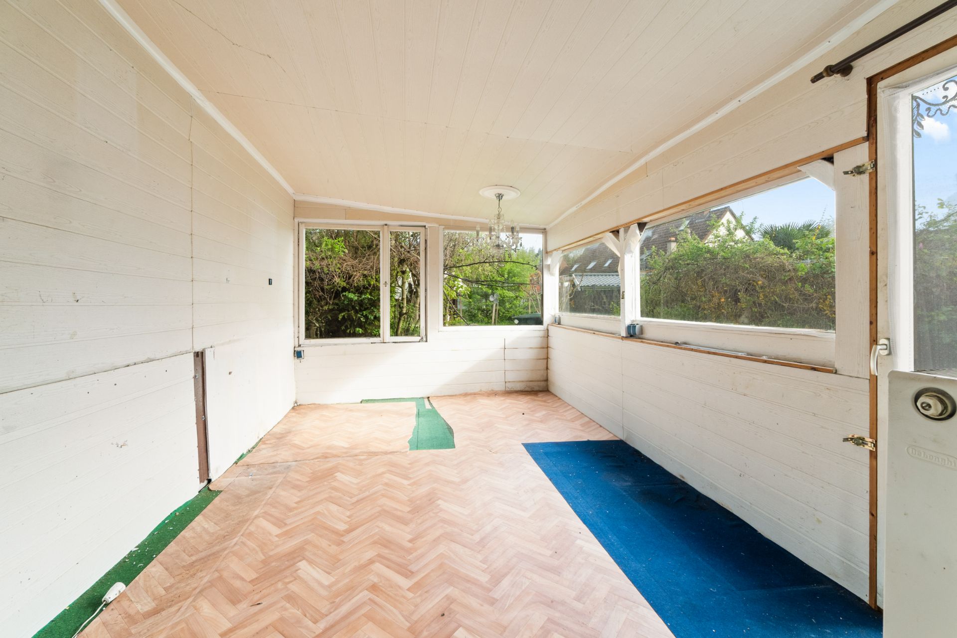 Heller Verandaraum mit Holzverkleidung und großen Fenstern, teilweise abgenutzter Bodenbelag.