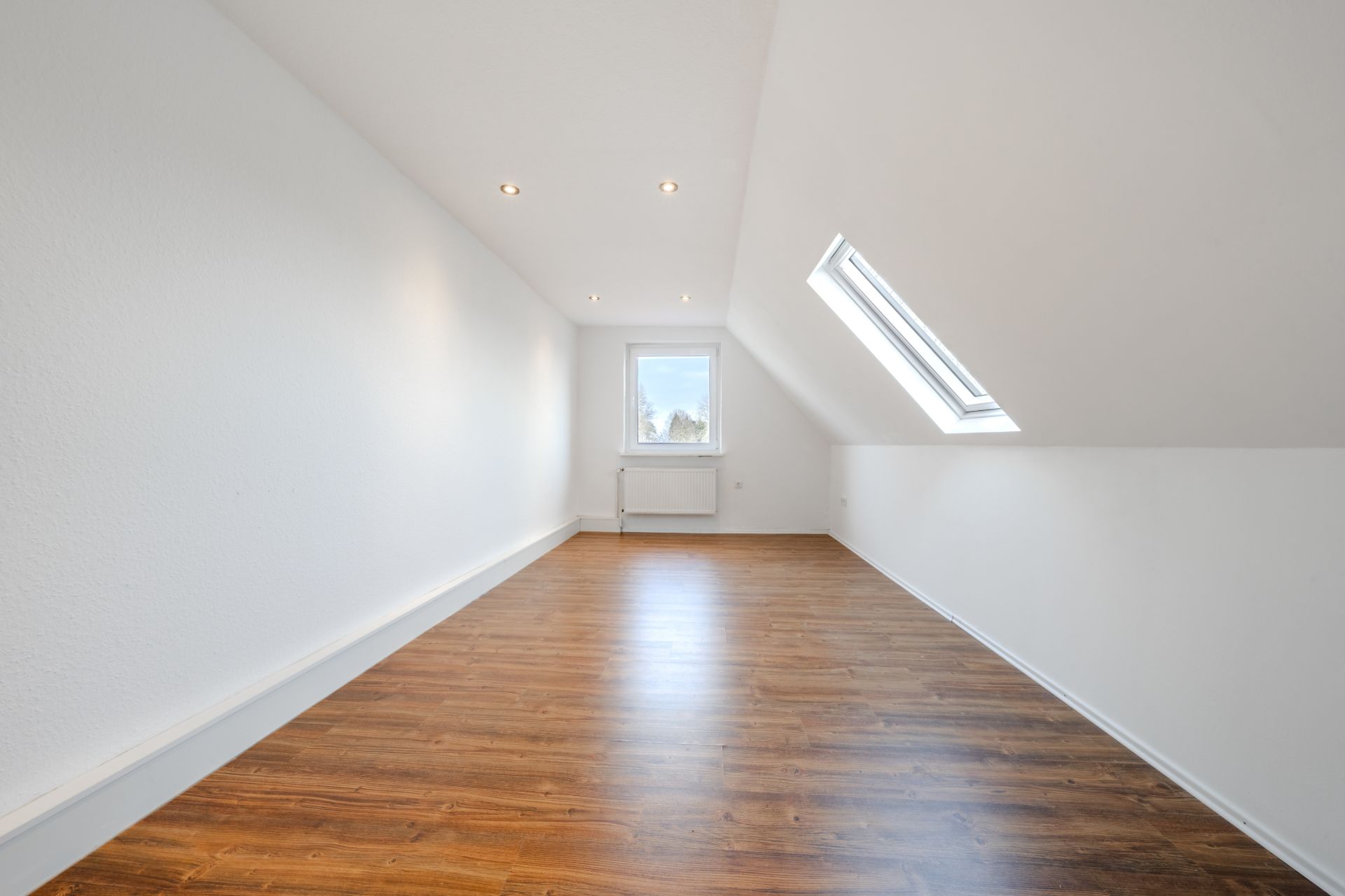 Heller Raum mit schräger Decke, Dachfenster, Holzfußboden und Fenster am Ende.