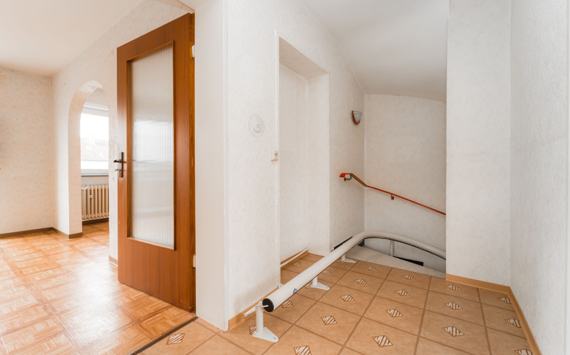 Heller Flur mit geöffneter Holztür, gefliestem Boden und Treppe mit Treppenlift.