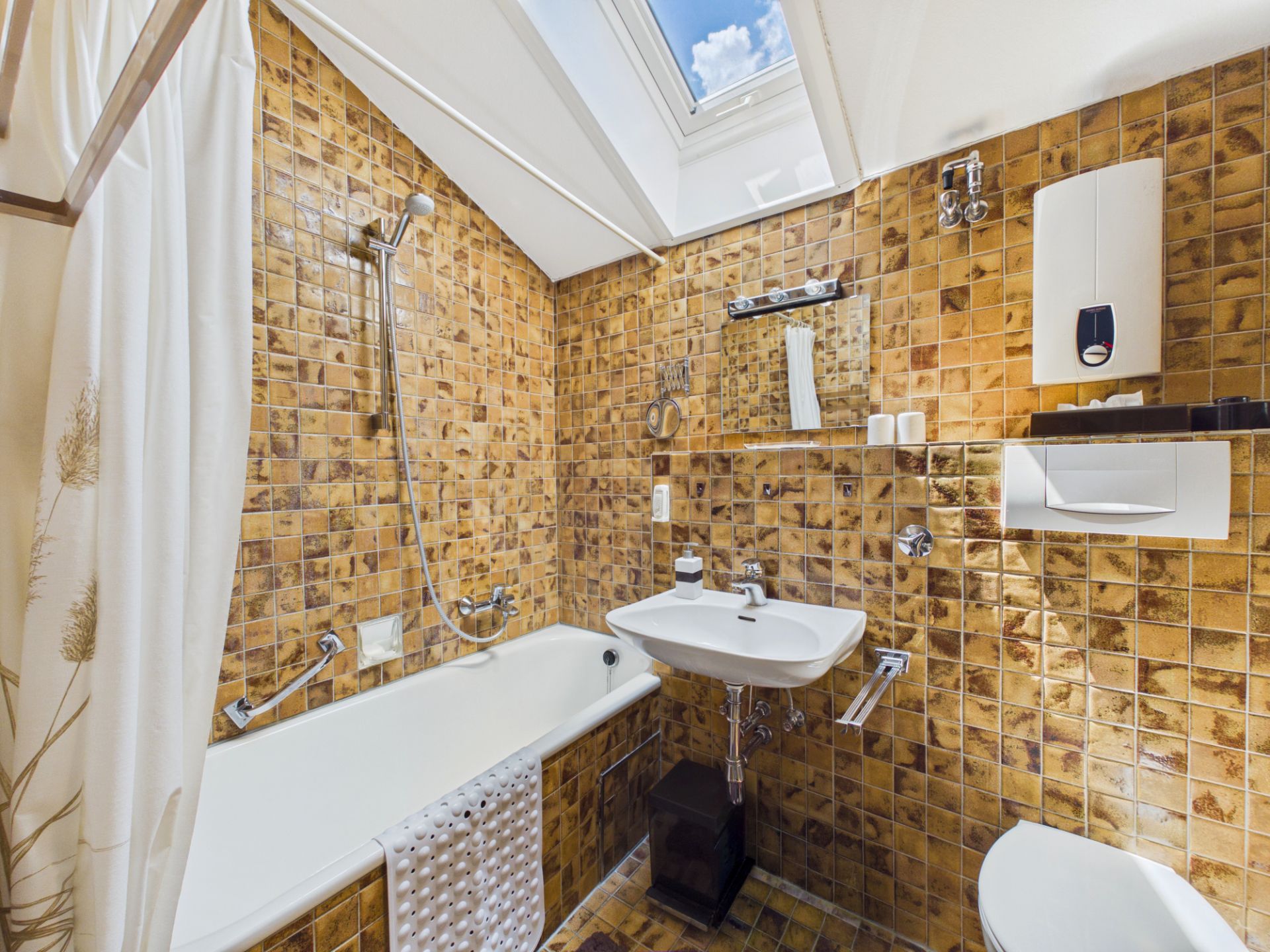 Kleines Badezimmer mit braunen Fliesen, Badewanne, Waschbecken, Toilette und Dachfenster.
