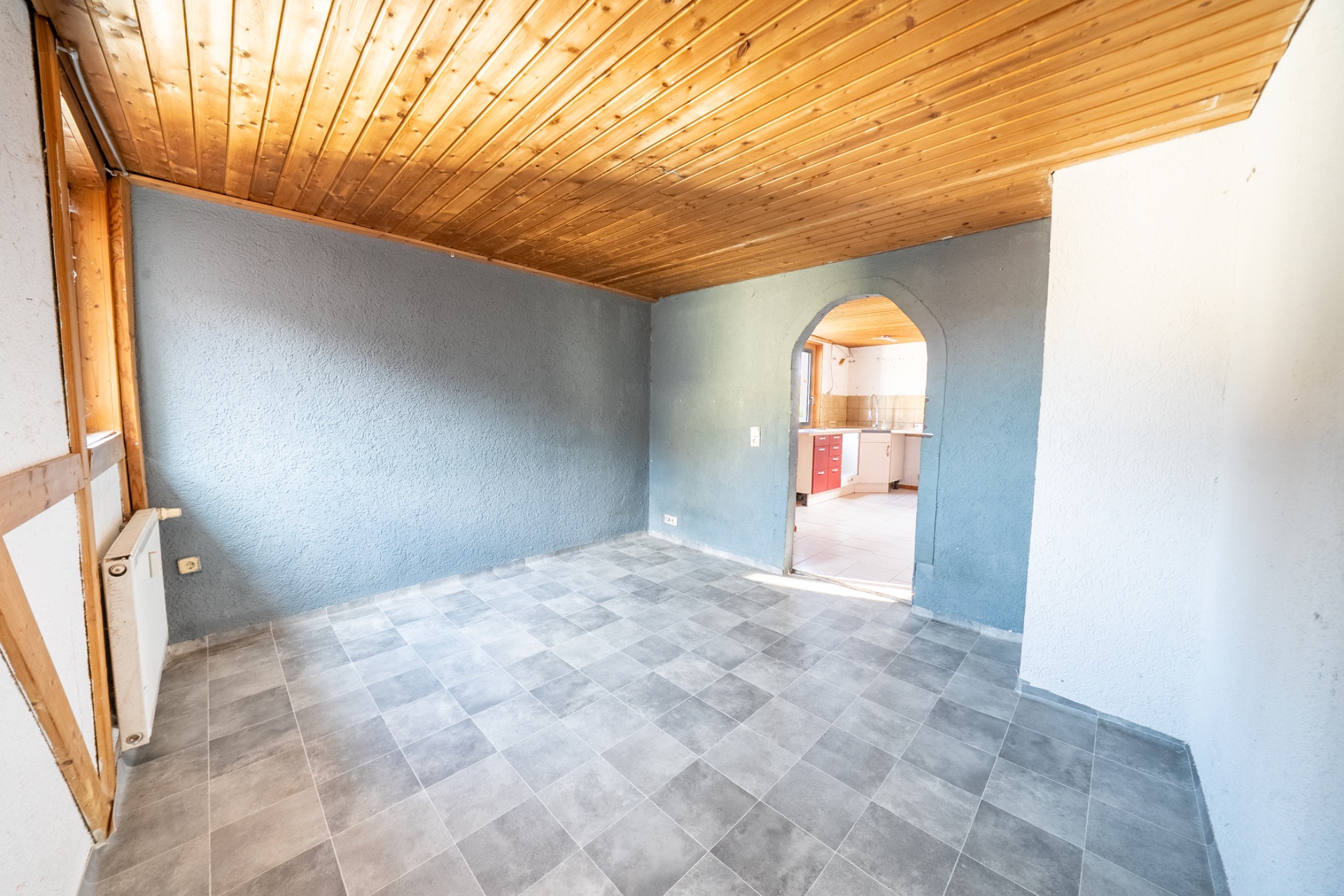 Leerer Raum mit grauem Boden, blauer Wand, Holzdecke und Durchgang zur Küche.