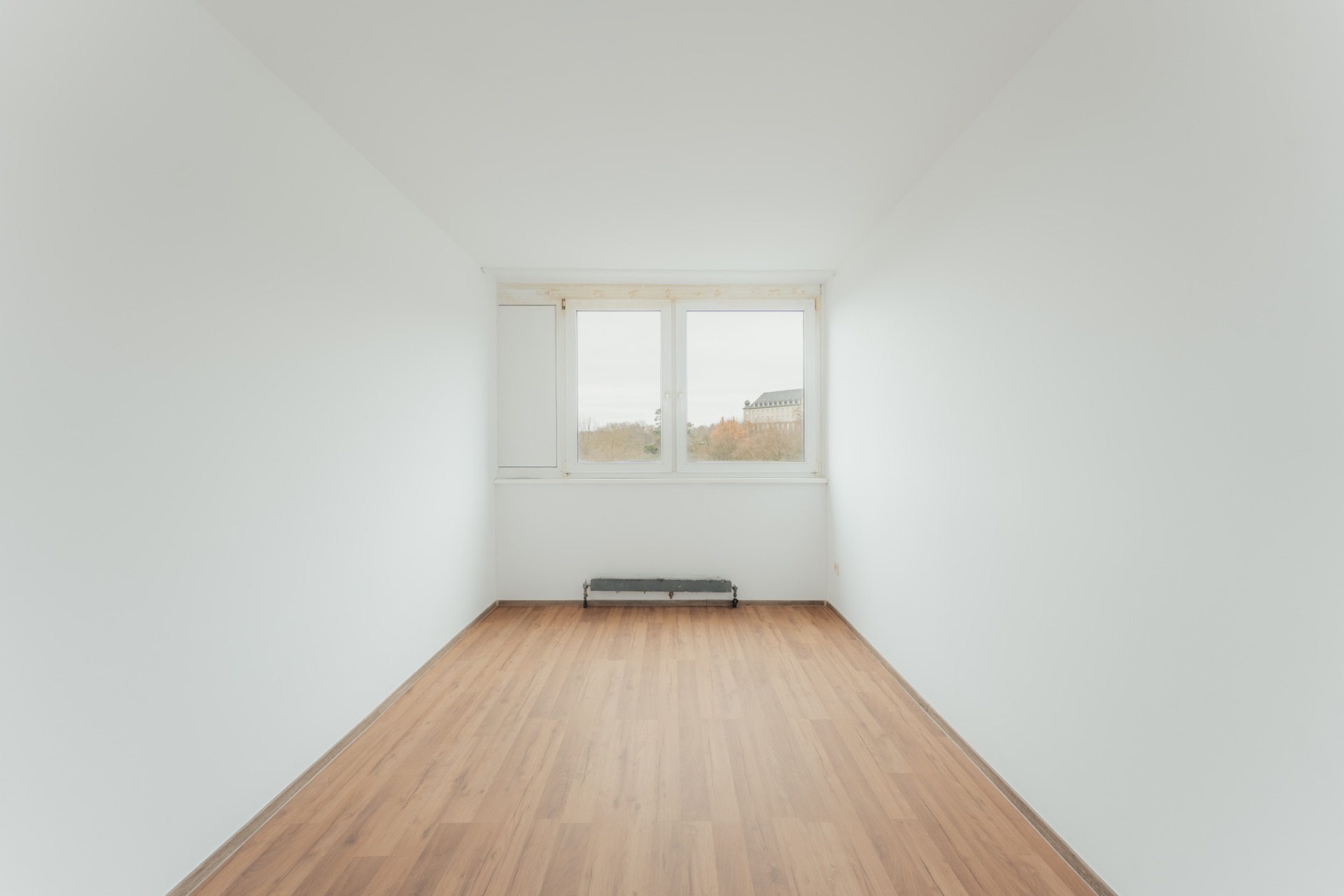 Leerer, heller Raum mit Holzfußboden und großem Fenster, weiße Wände.