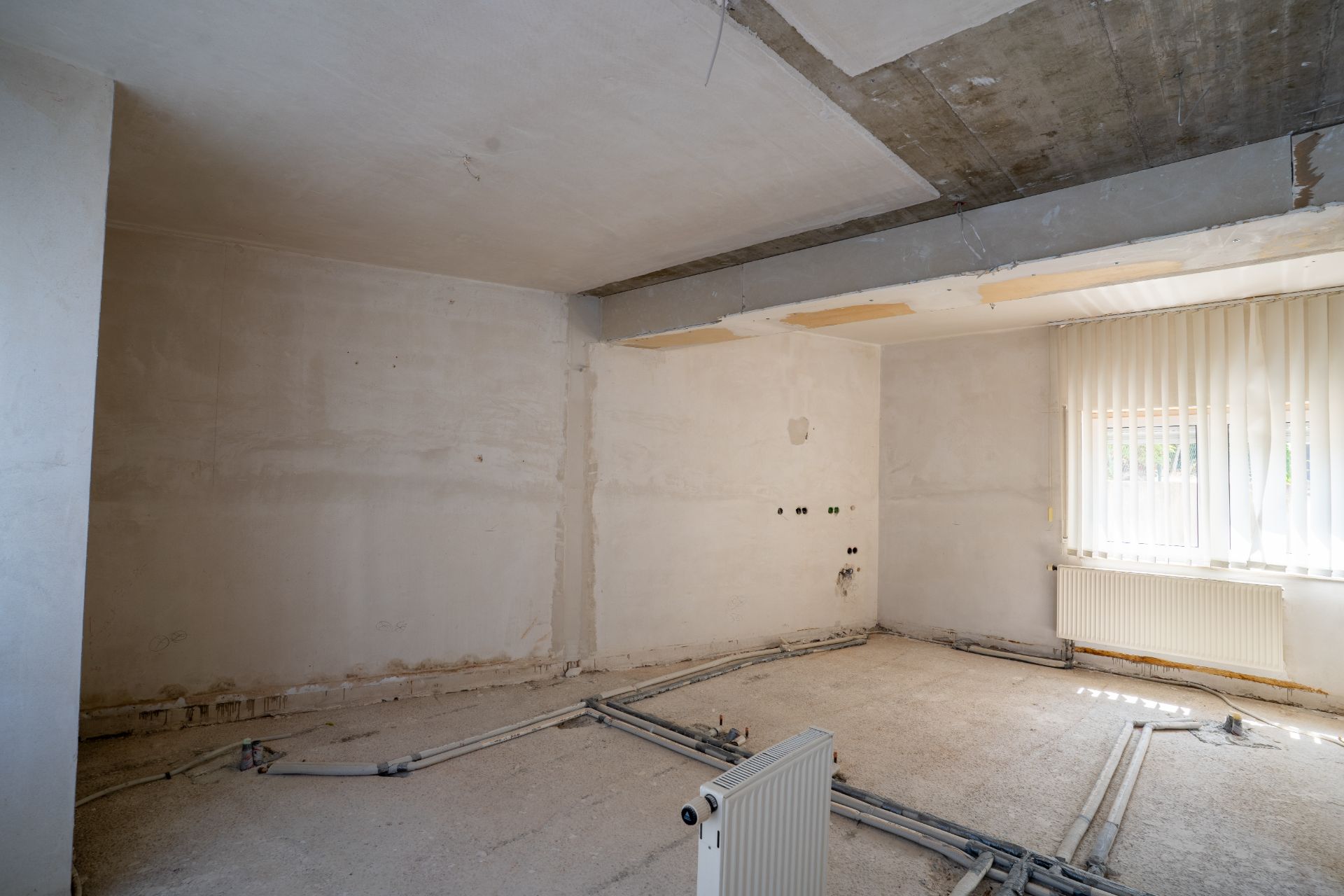 Leerer Raum mit sichtbaren Rohren am Boden, weiß gestrichenen Wänden und einem Fenster mit Lamellenvorhang.