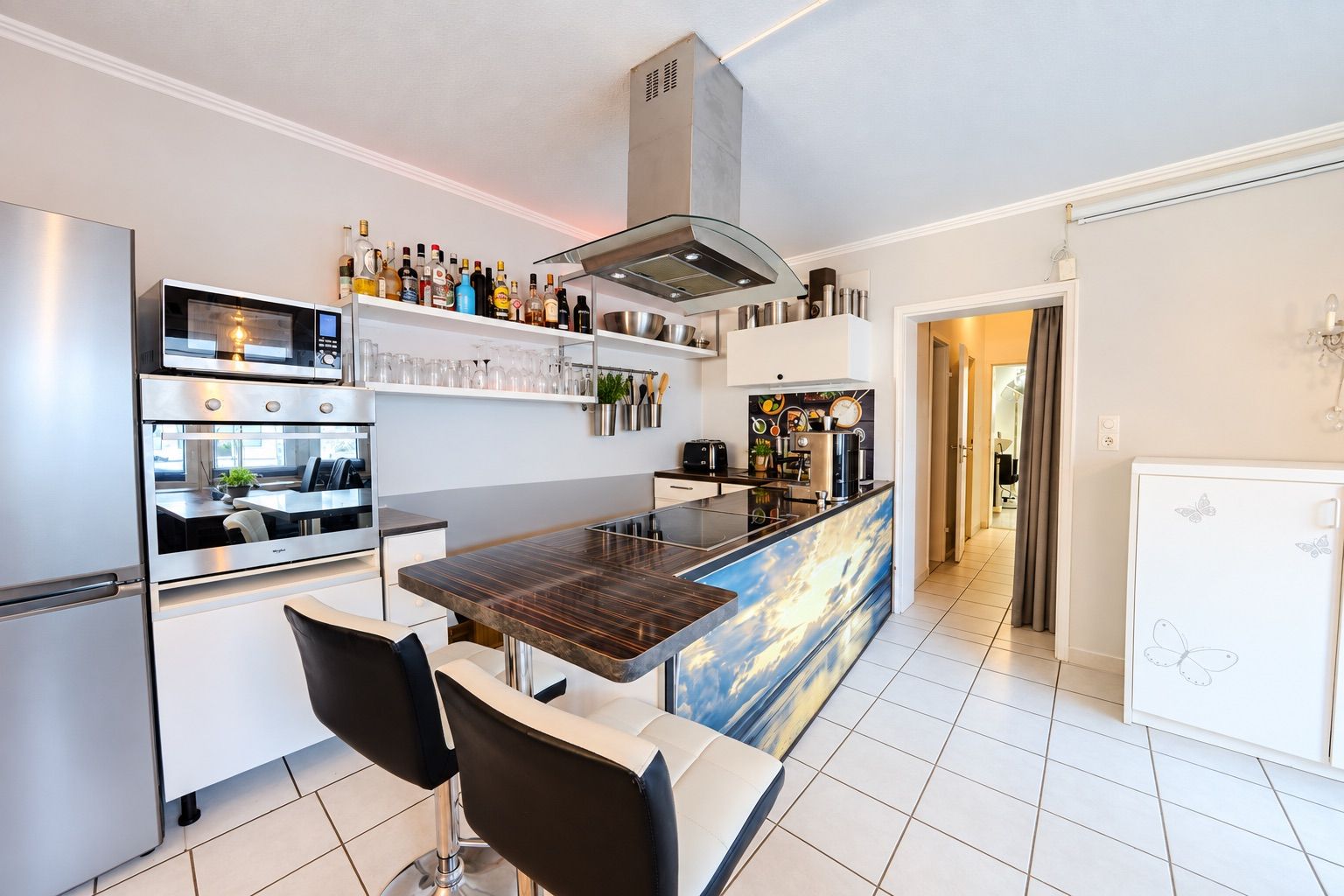 Moderne Küche mit Kochinsel, zwei Barhockern, Kühlschrank und Einbaugeräten auf Fliesenboden.