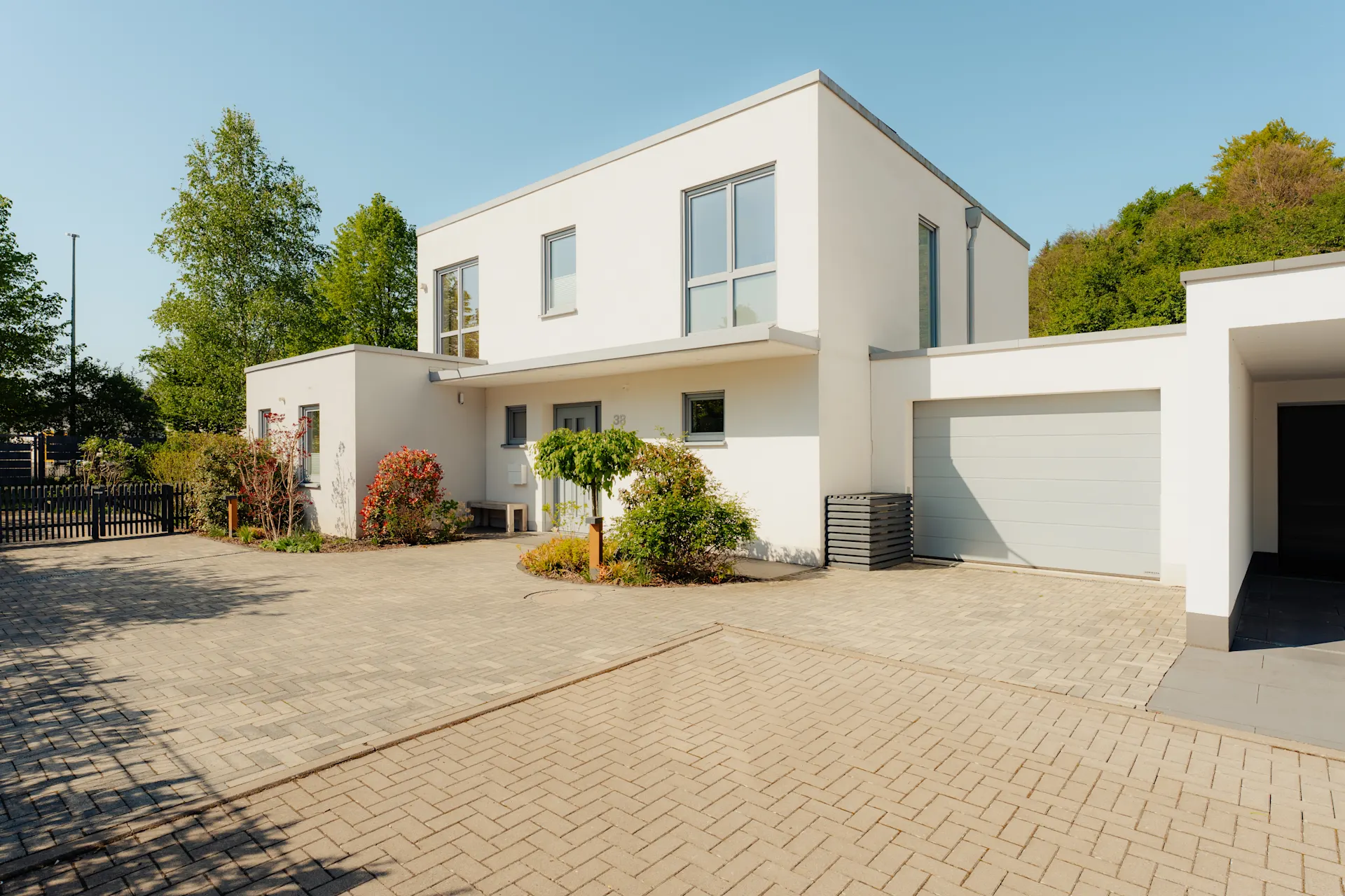 Modernes, weißes Wohnhaus mit Flachdach, großen Fenstern und Garage, umgeben von Bäumen und Pflanzen.