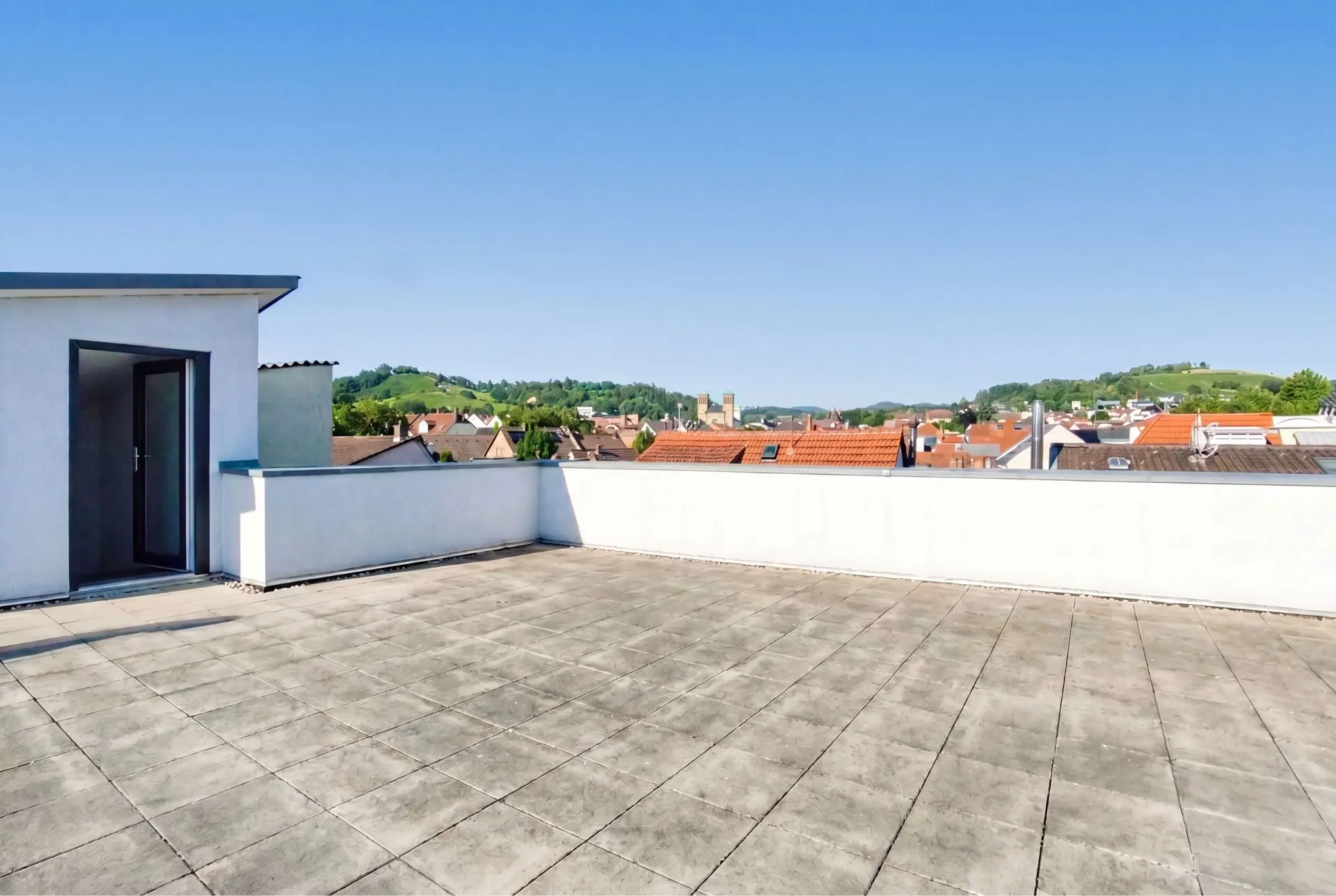 Große Dachterrasse mit Fliesenboden und Blick auf Wohnhäuser und Hügel.