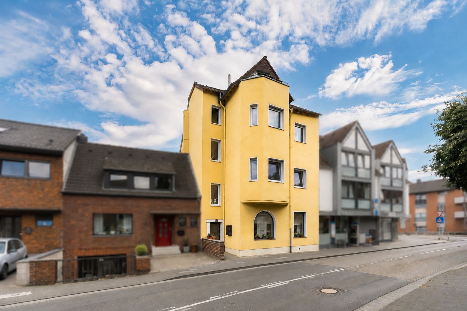 Gelbes dreistöckiges Wohnhaus mit Spitzdach an einer ruhigen Straße, daneben weitere Wohnhäuser.
