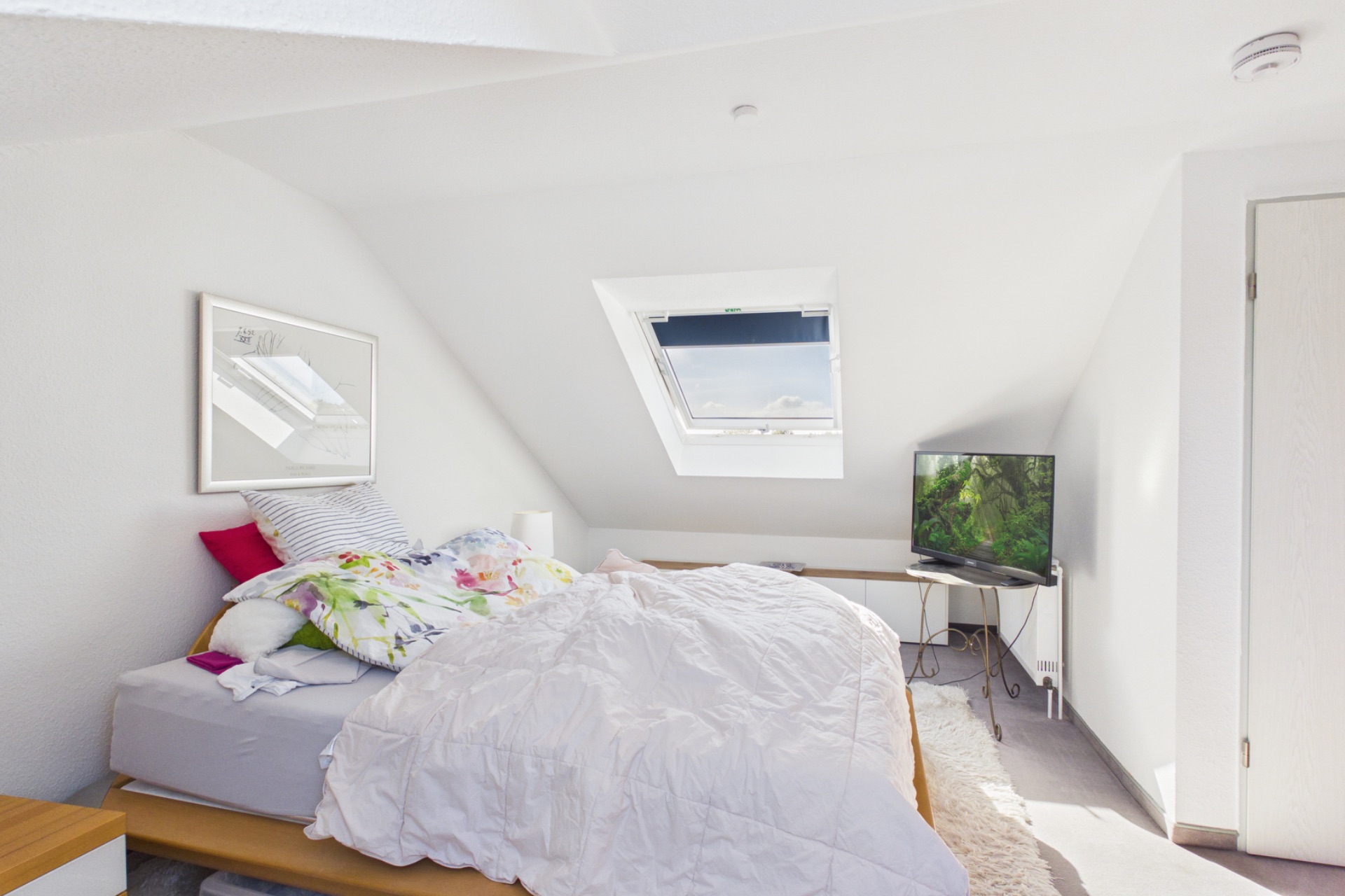 Helles Schlafzimmer mit schräger Decke, Bett, Fenster, Fernseher und Kommode.