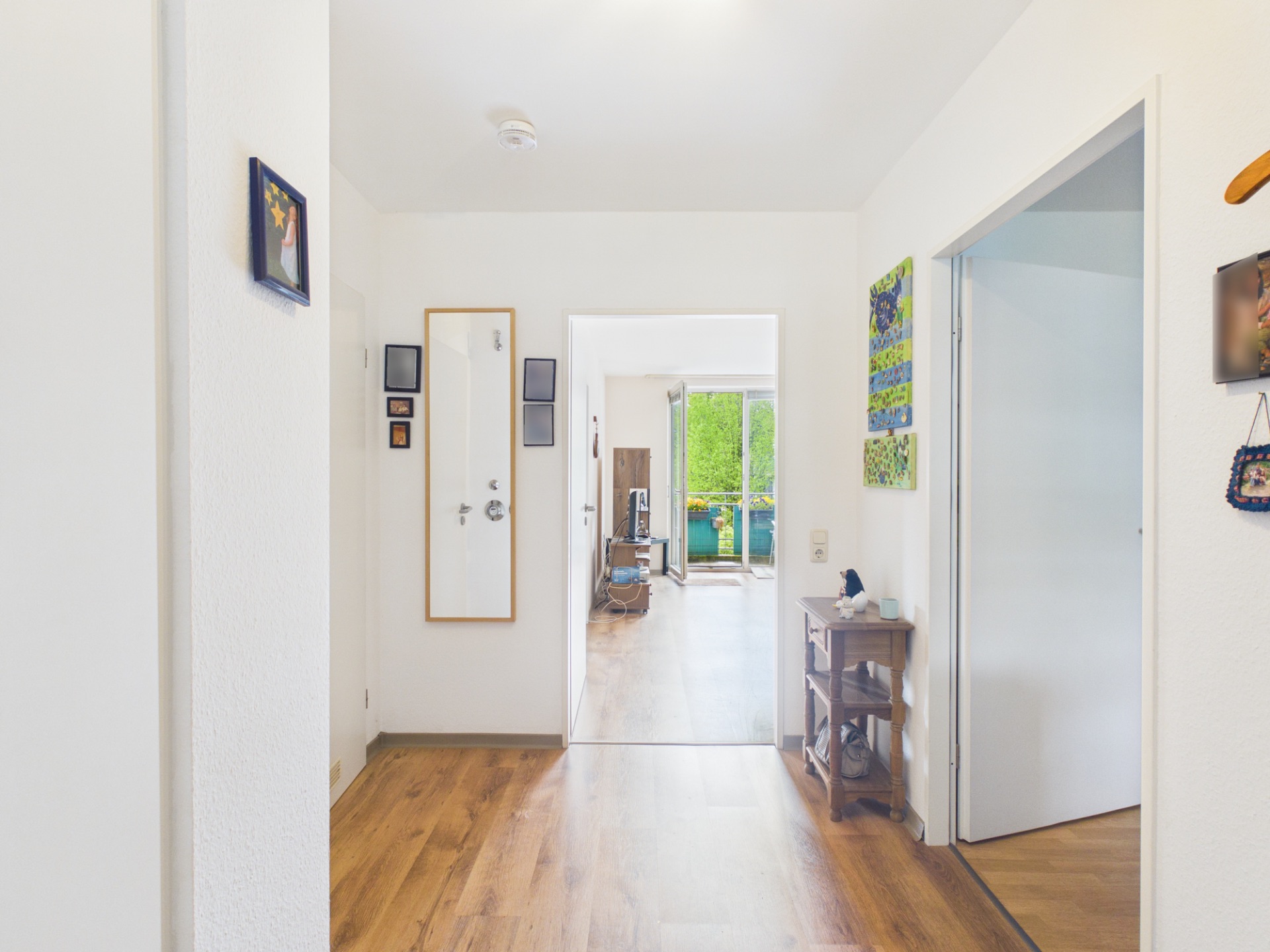 Heller Flur mit Holzboden, Wandbildern und Blick ins Wohnzimmer mit Balkon.