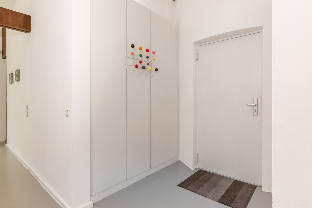 Heller Flur mit weißer Tür, Einbauschrank und bunter Garderobe, graue Fußmatte am Boden.