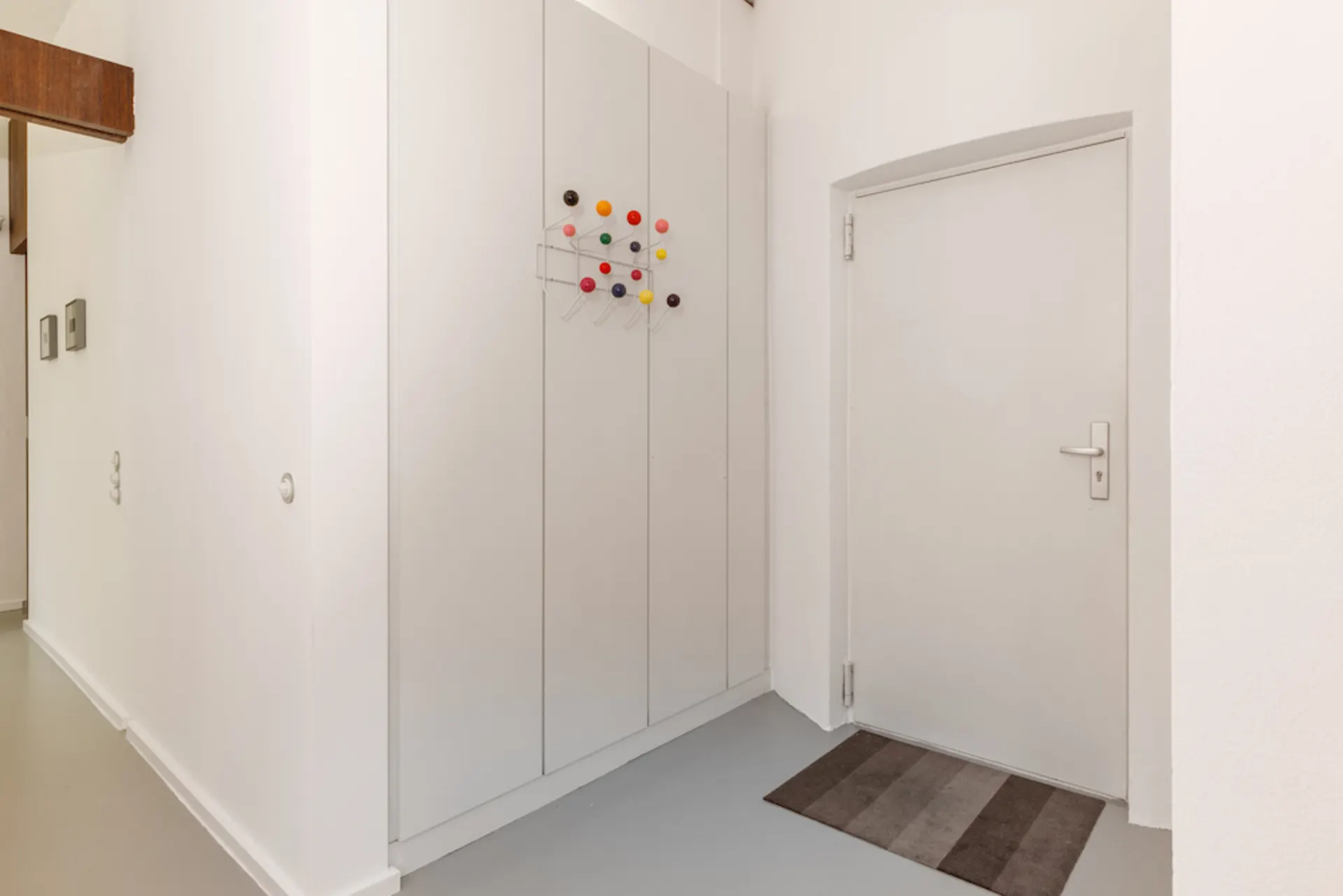 Heller Flur mit weißer Tür, Einbauschrank und bunter Garderobe, graue Fußmatte am Boden.