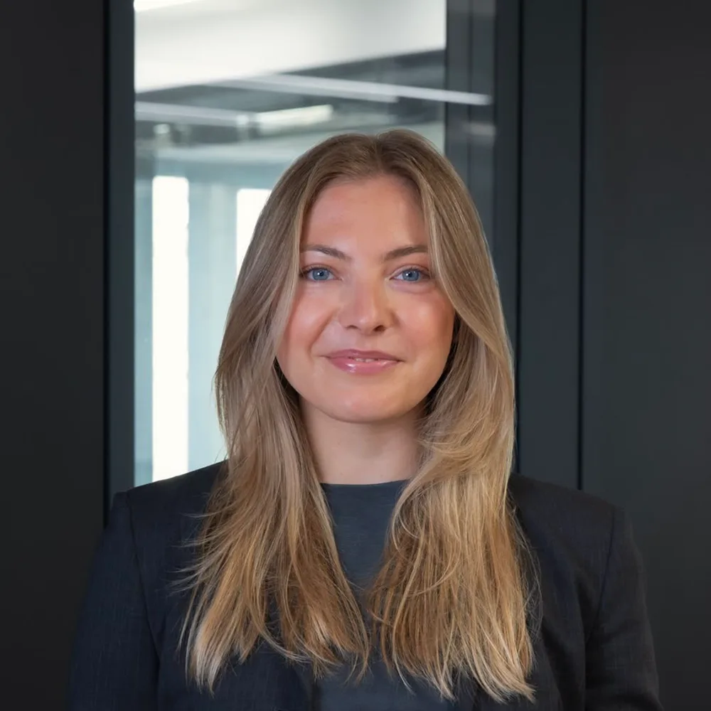 Eine Frau mit langen blonden Haaren steht in einem dunklen Anzug vor einer Glaswand.