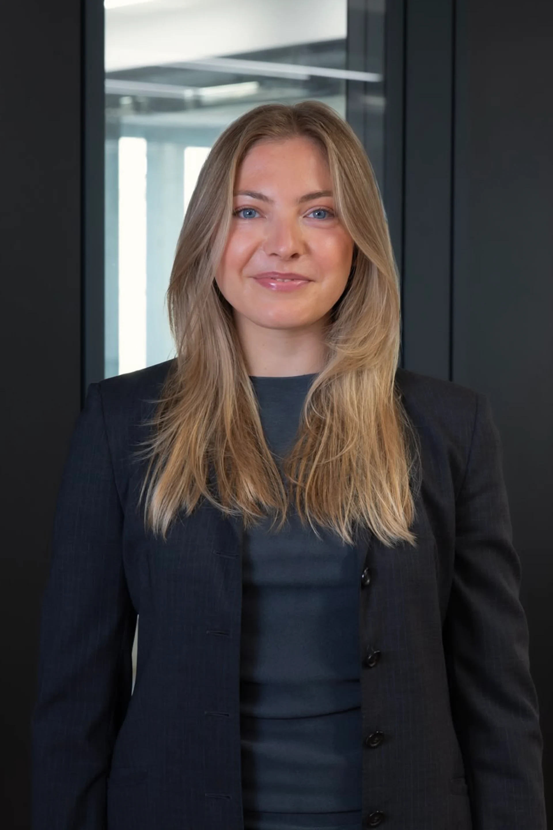 Eine Frau mit langen blonden Haaren steht in einem dunklen Anzug vor einer Glaswand.