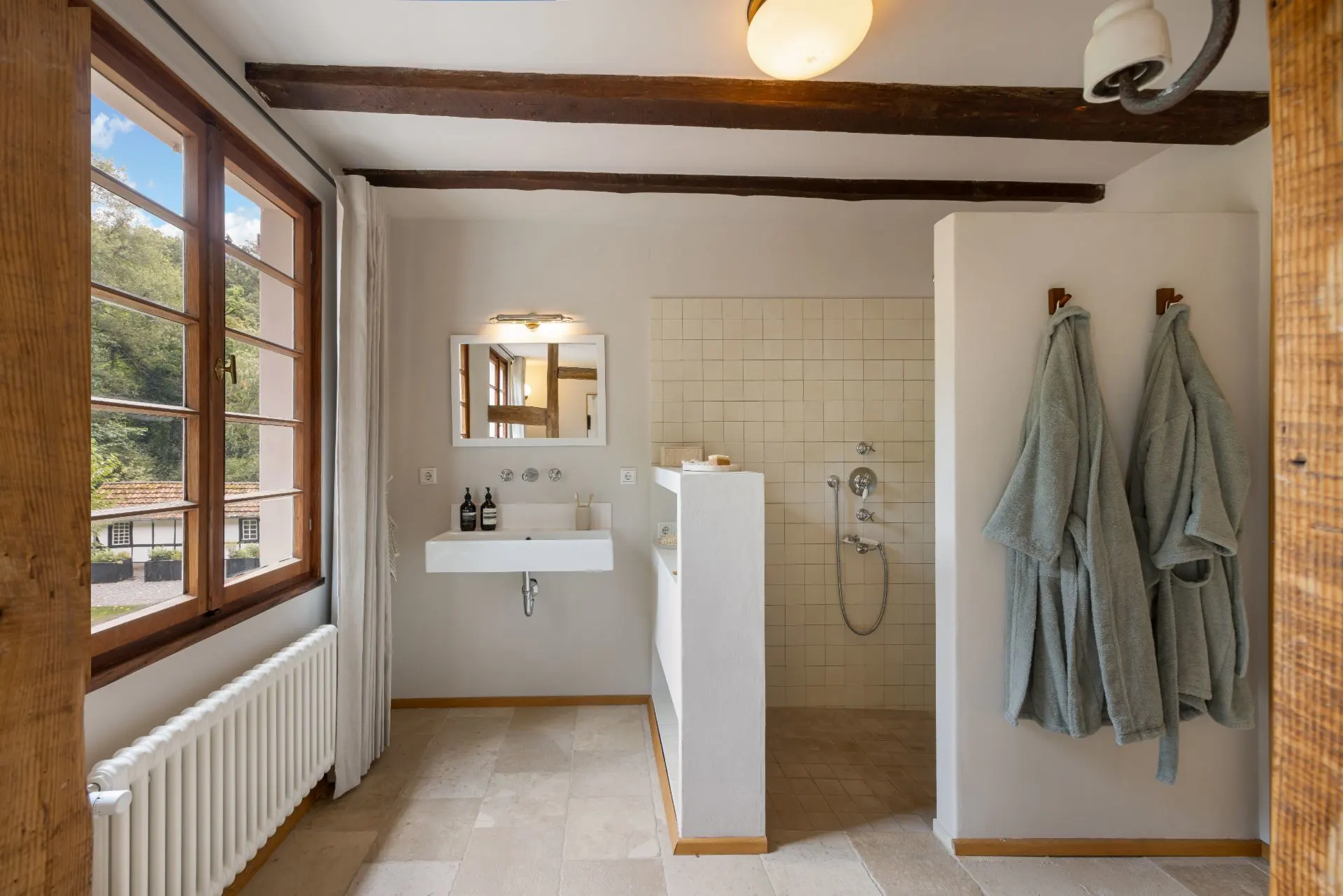 Helles Badezimmer mit großem Fenster, Waschbecken, Spiegel, offener Dusche und zwei Bademänteln an der Wand.