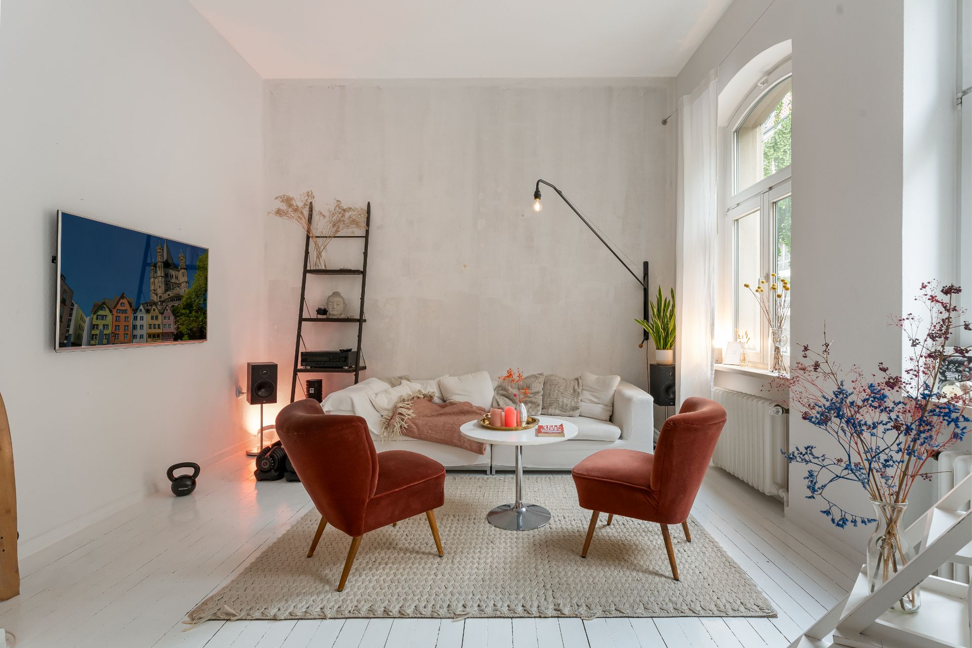 Helles Wohnzimmer mit weißem Sofa, zwei roten Sesseln, Teppich, Fernseher und großen Fenstern.