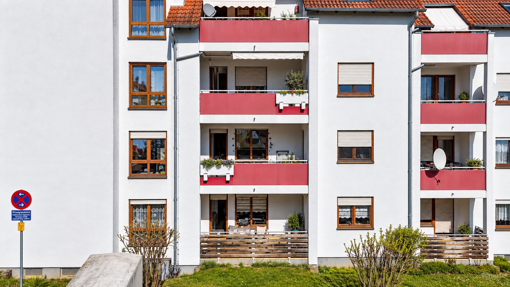 Mehrfamilienhaus mit roten Balkonen und mehreren Fenstern, davor grüne Wiese.