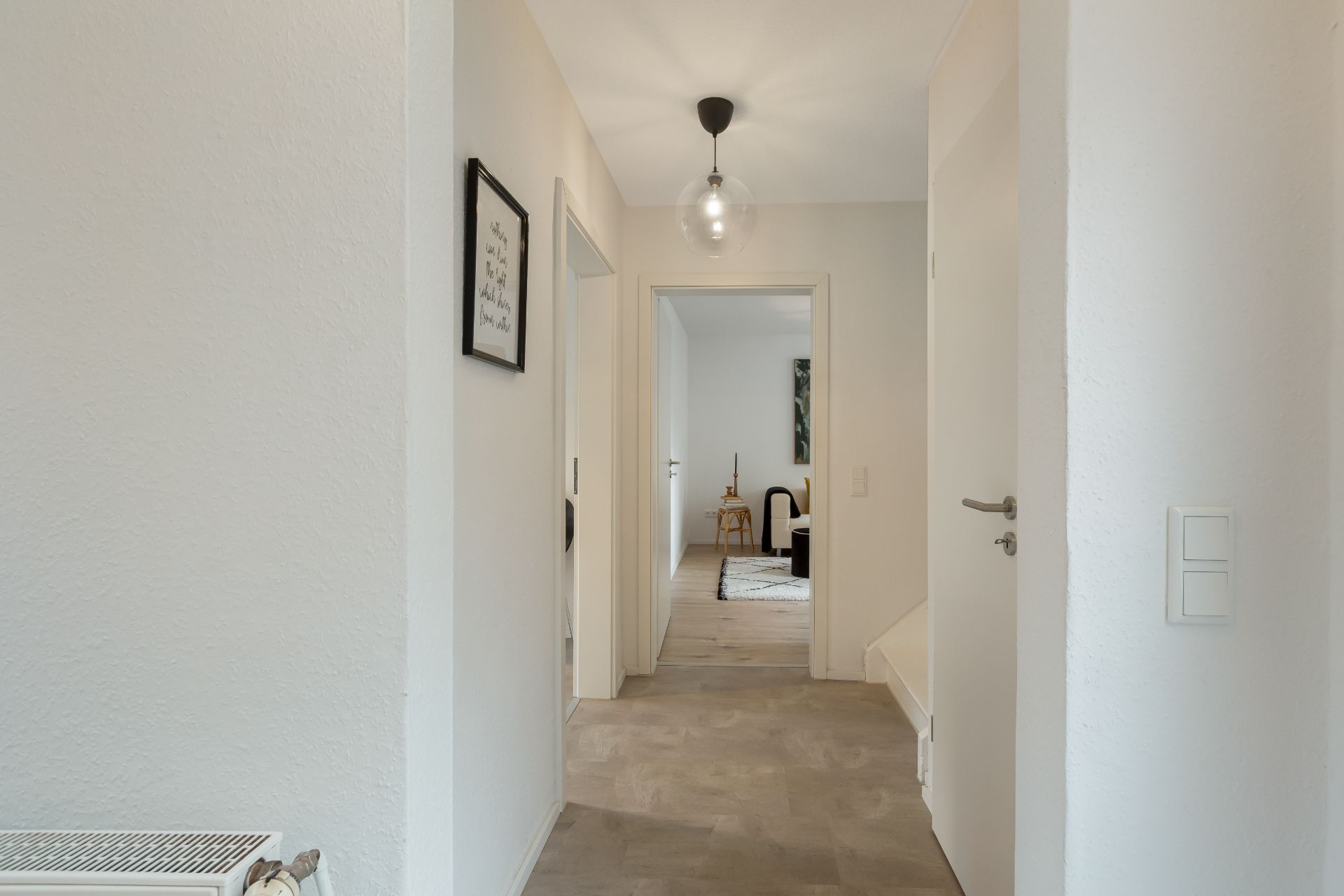 Heller Flur mit offener Tür zu einem Zimmer, Wandleuchte und gerahmtem Bild an der Wand.