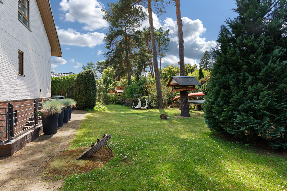 Garten mit grüner Wiese, Bäumen, Schaukel und Vogelhaus neben einem weißen Wohnhaus.