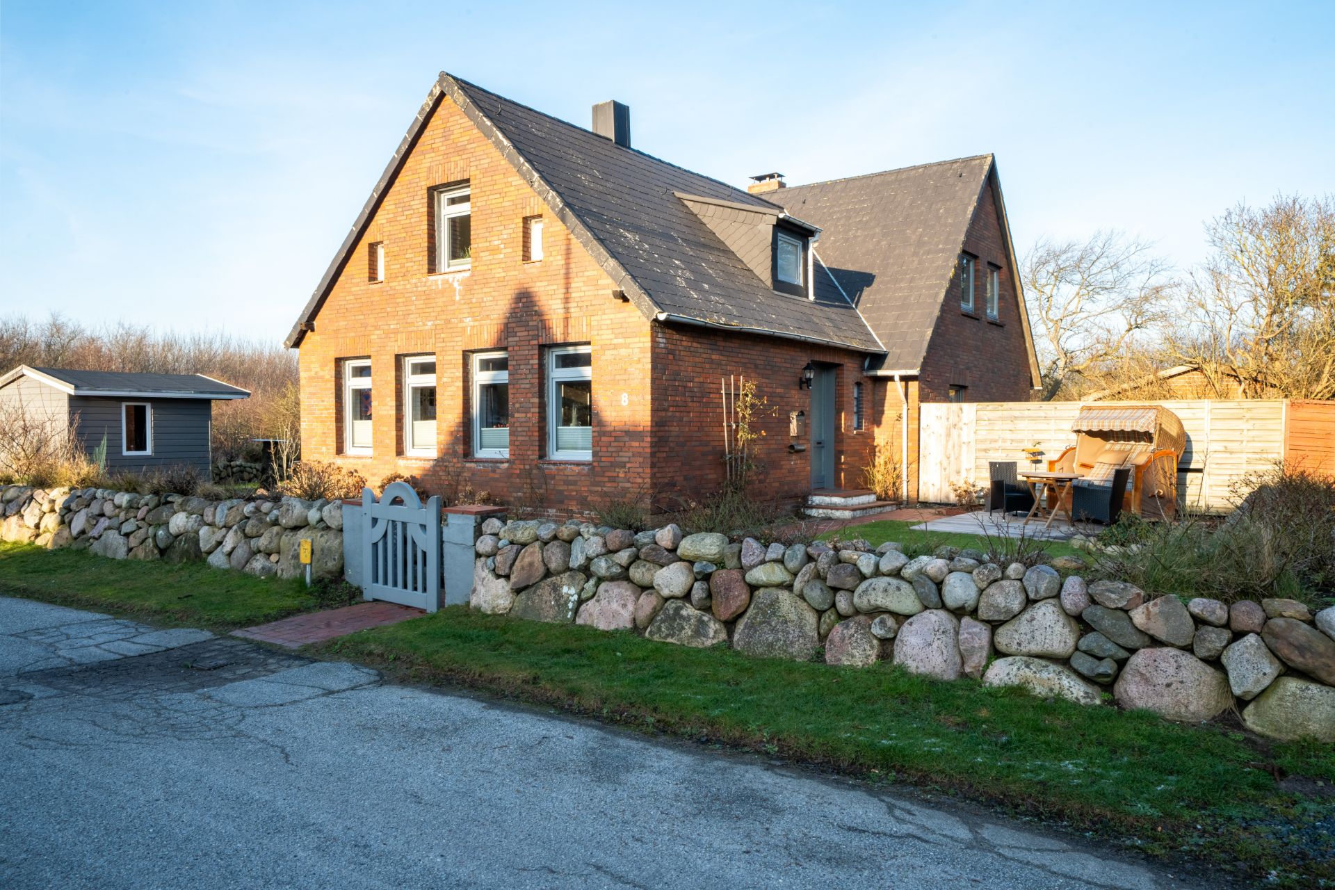 Backsteinhaus mit dunklem Dach, Steinmauer, Garten und Terrasse mit Gartenmöbeln.