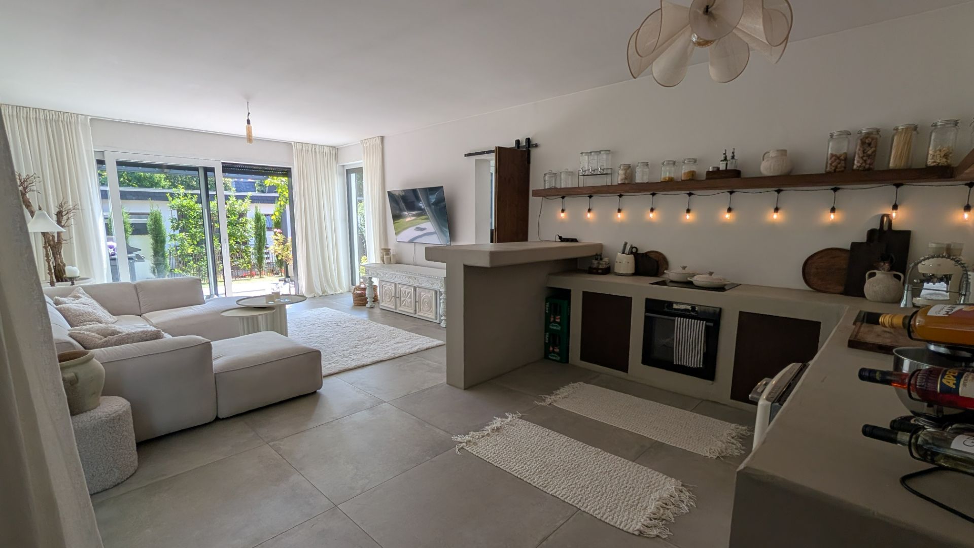 Helles Wohnzimmer mit offener Küche, Sofa, Fernseher, großem Fenster und moderner Einrichtung.