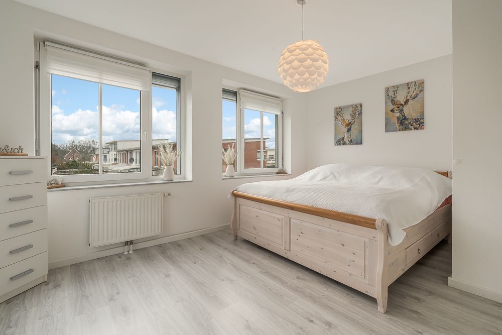 Helles Schlafzimmer mit großem Bett, drei Fenstern, Kommode und zwei Wandbildern.