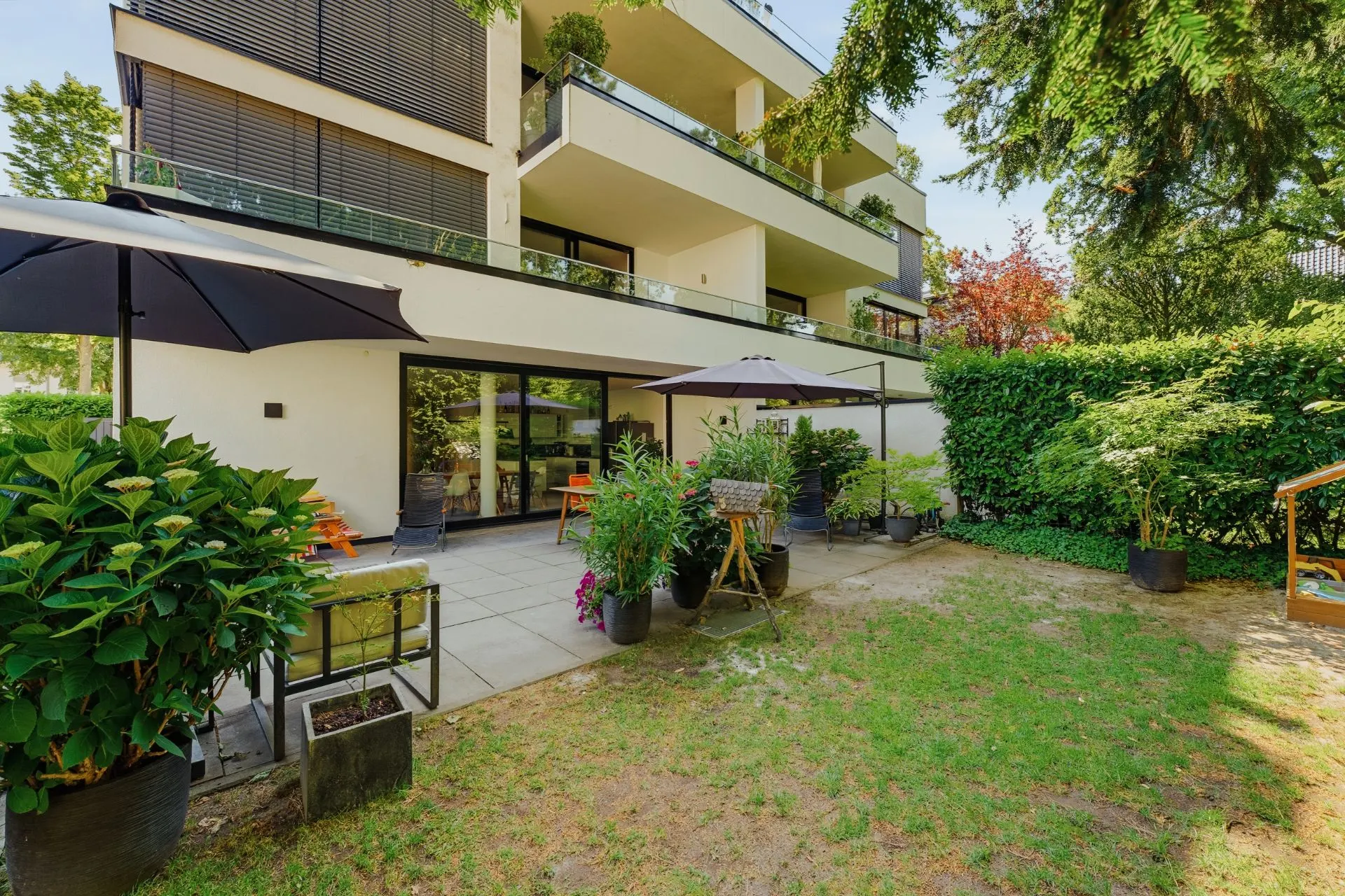Moderne Terrasse mit Garten, Sonnenschirmen, Pflanzen und Sitzbereich vor einem Wohnhaus.