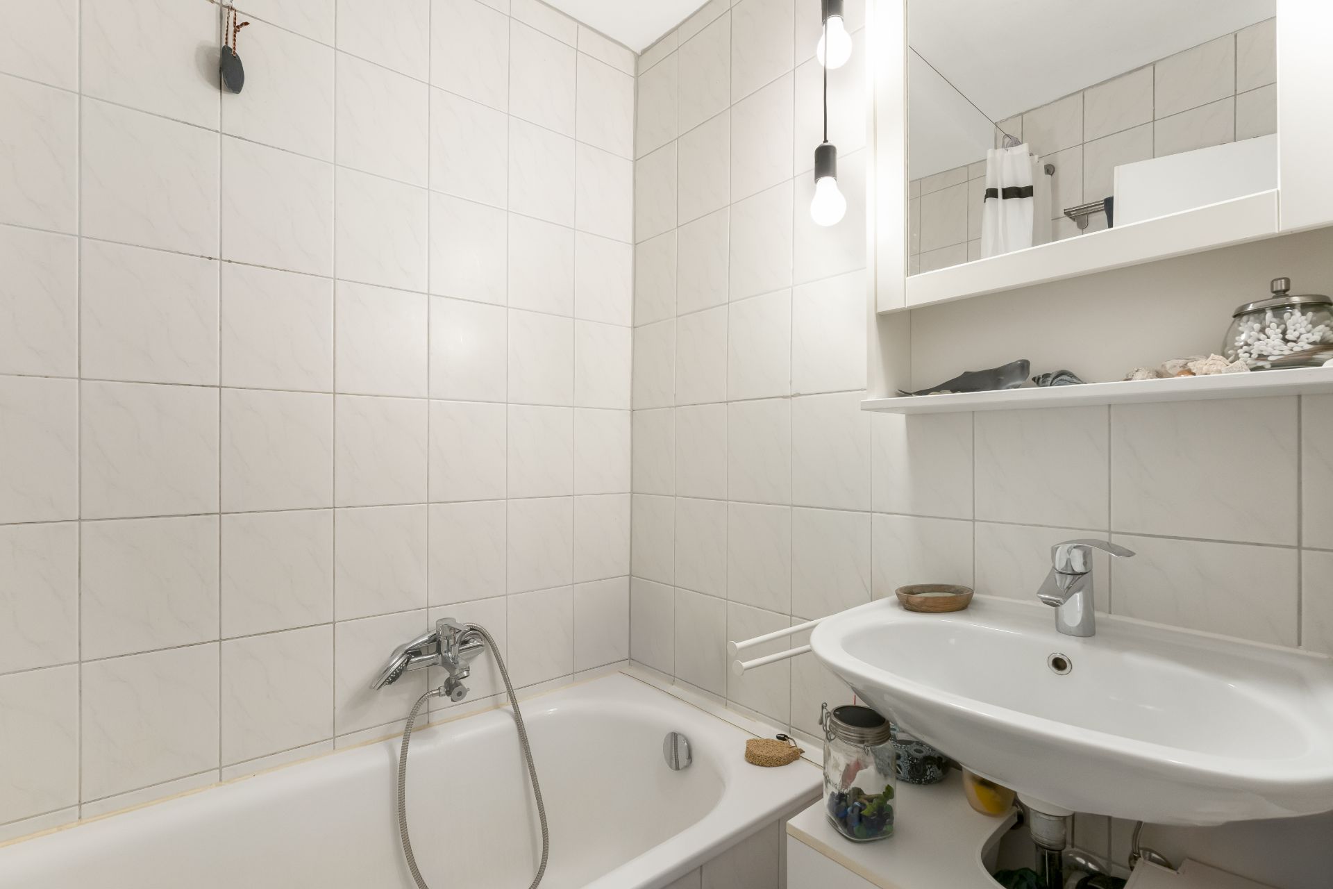 Helles Badezimmer mit Badewanne, Waschbecken, Wandspiegel und Regal mit Gegenständen.