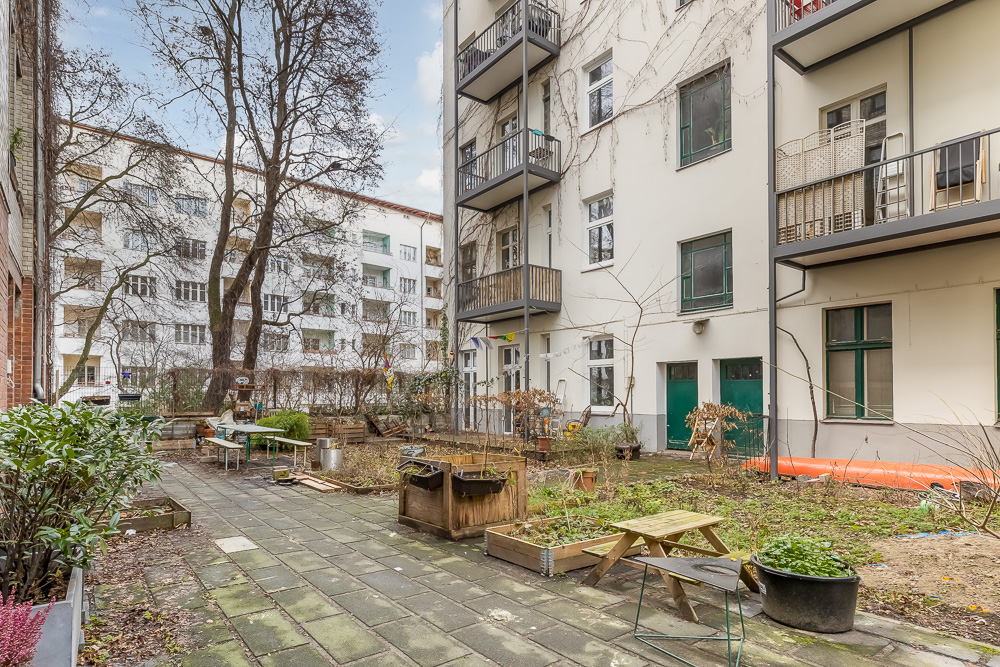 Hinterhof mit Pflasterweg, Beeten, Bäumen, Tischen und Mehrfamilienhaus mit Balkonen.