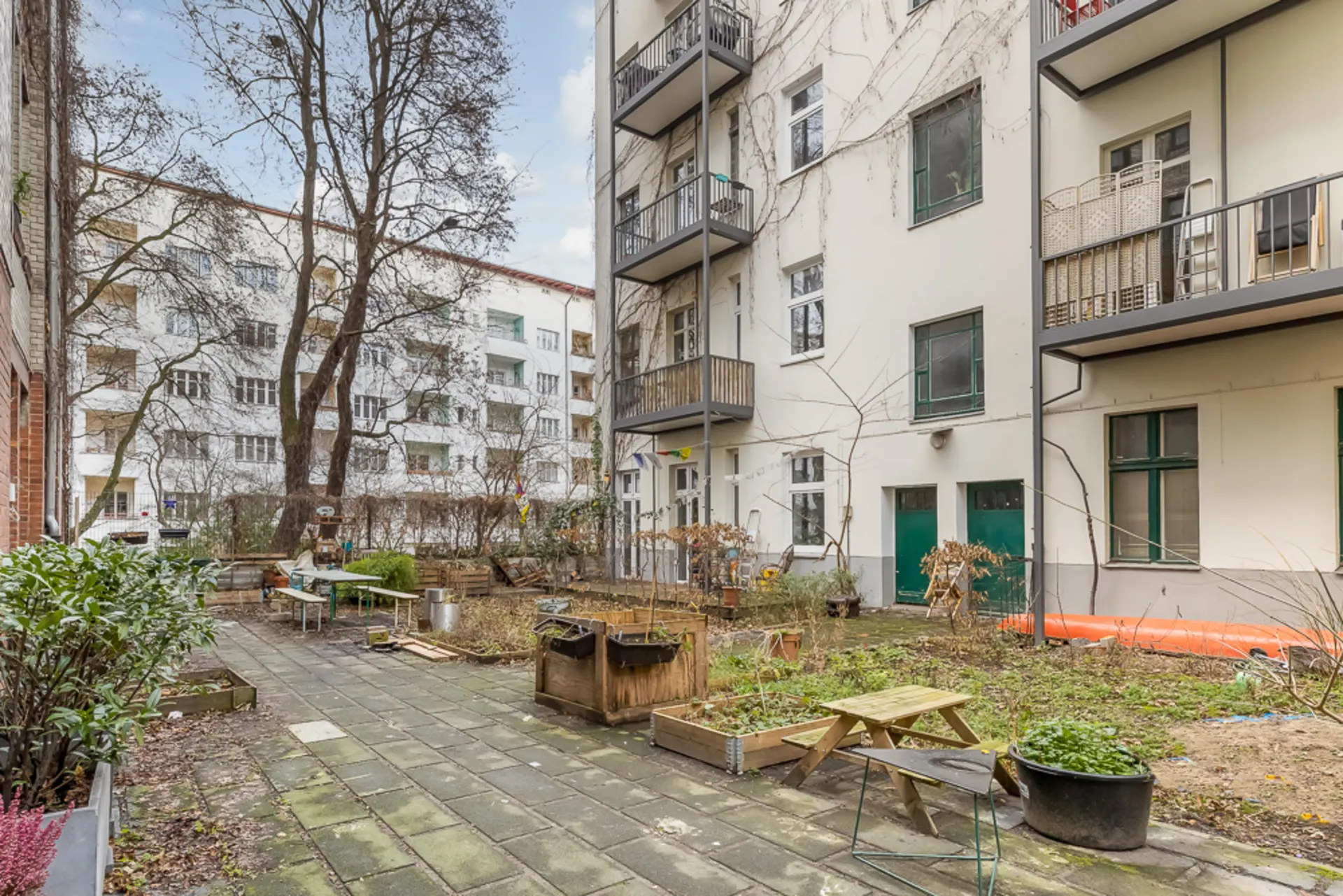 Hinterhof mit Pflasterweg, Beeten, Bäumen, Tischen und Mehrfamilienhaus mit Balkonen.