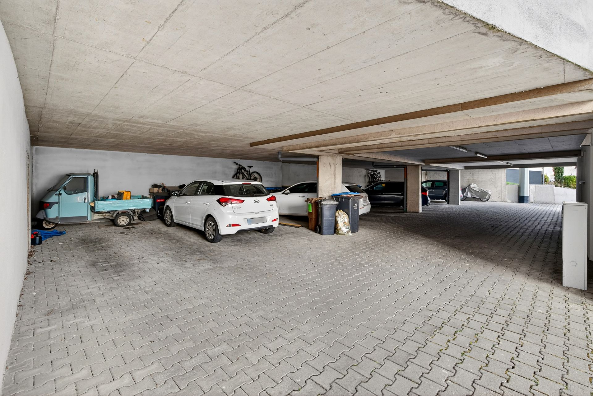 Überdachte Parkfläche mit mehreren geparkten Autos, Mülltonnen und einem kleinen blauen Fahrzeug.
