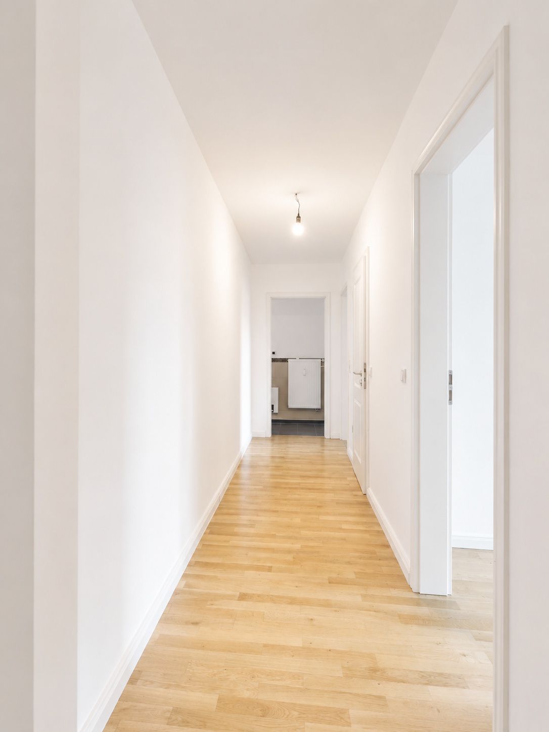 Heller Flur mit Holzfußboden, weißen Wänden und offener Tür am Ende.