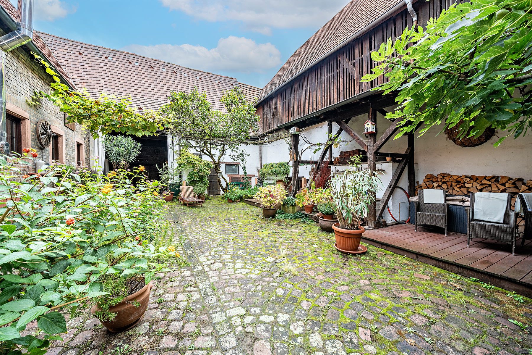 Gepflasterter Innenhof mit vielen Pflanzen, Gartenmöbeln und Holzlager unter einem Vordach.