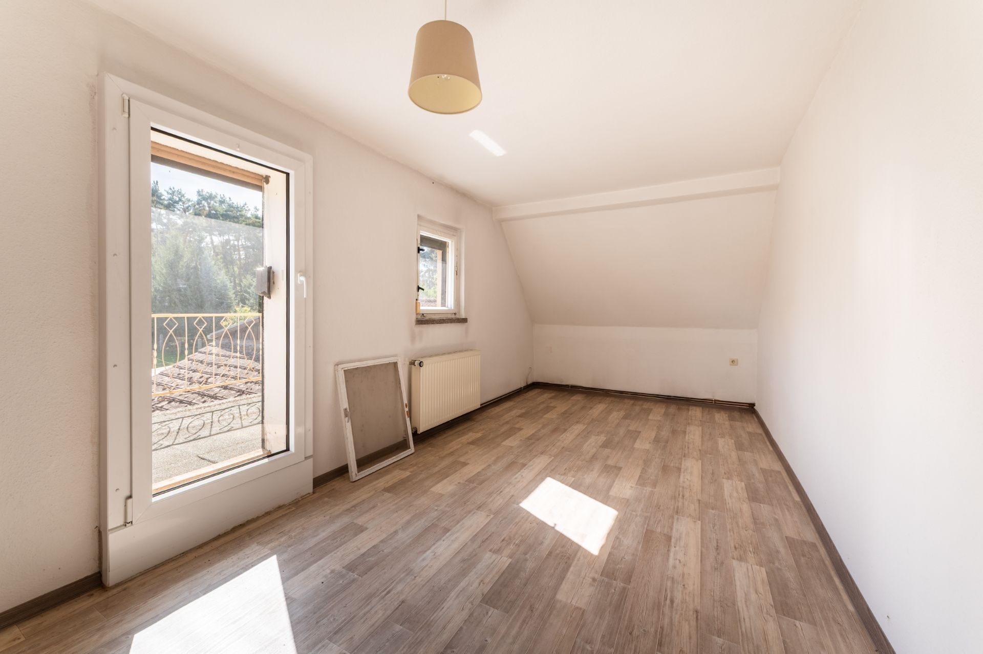 Leeres Zimmer mit Holzfußboden, Fenster, Tür zum Balkon und Hängelampe an der Decke.