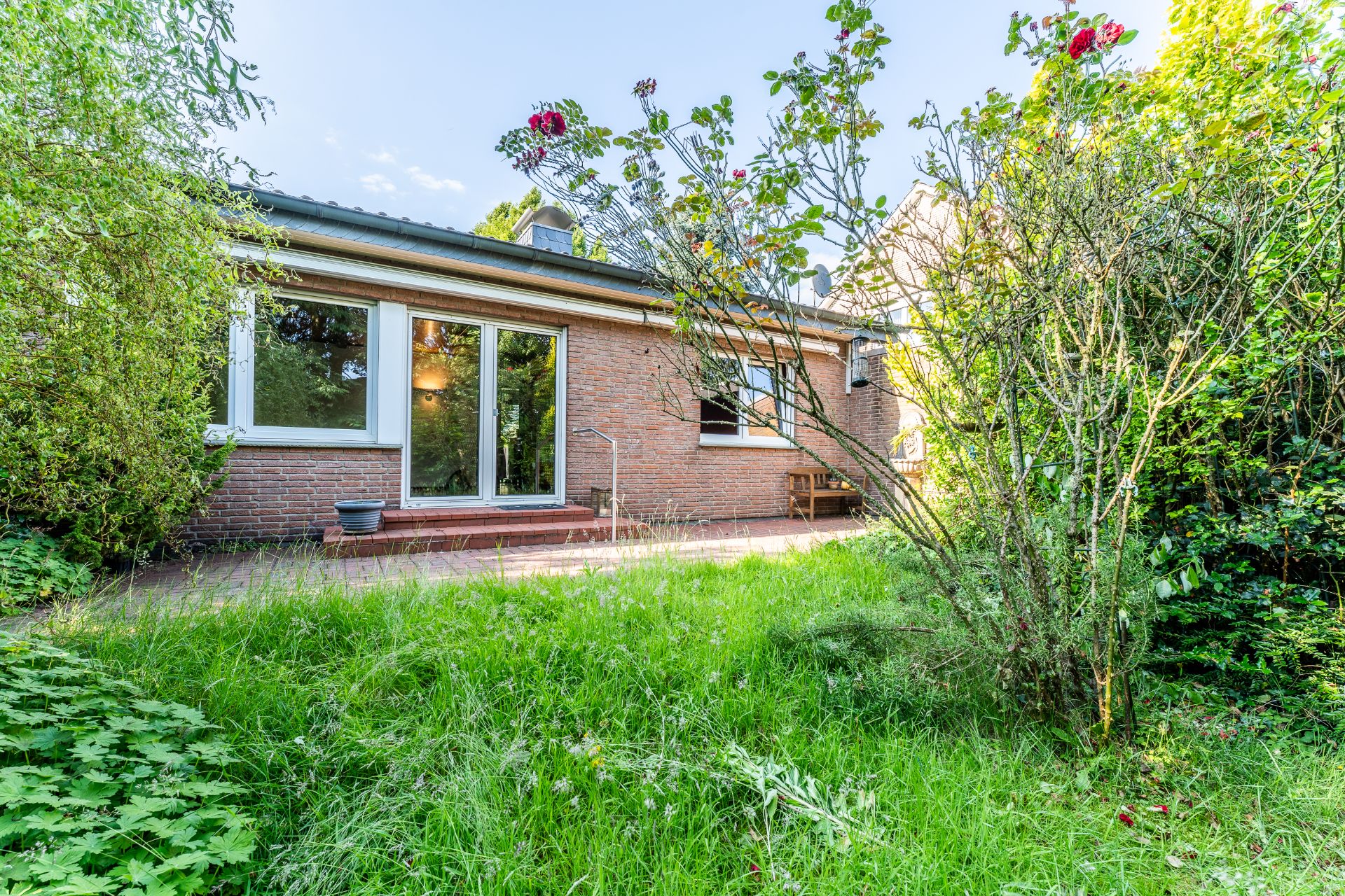 Backstein-Haus mit großen Fenstern, Terrasse und Garten mit Blumen und Sträuchern.