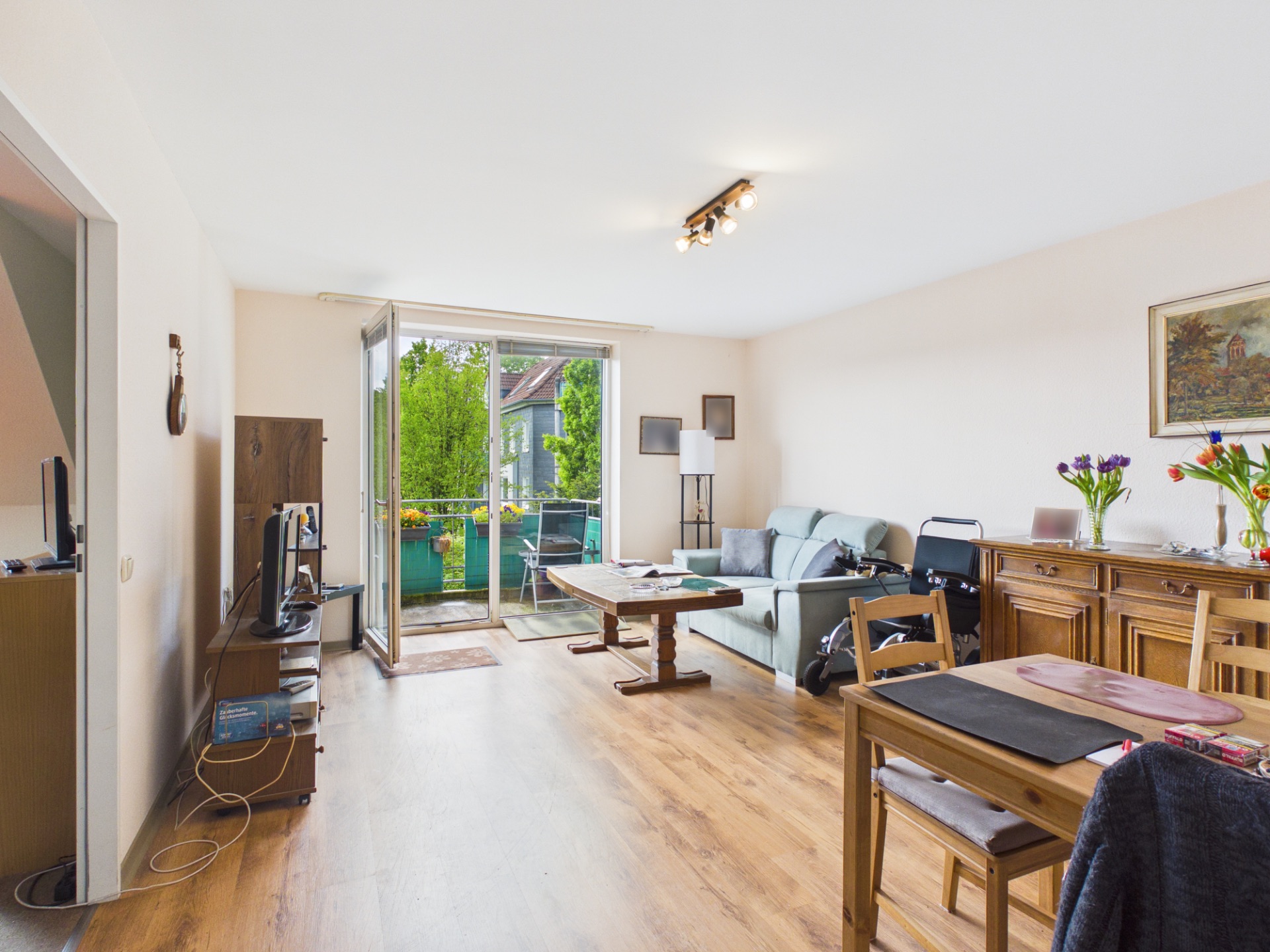 Helles Wohnzimmer mit Holzfußboden, Sofa, Esstisch, Sideboard und Zugang zum Balkon.