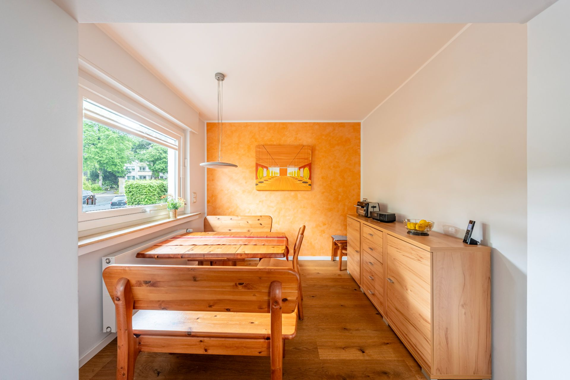 Esszimmer mit Holztisch, Bänken, Kommode und großem Fenster, Wand in Orange gestrichen.