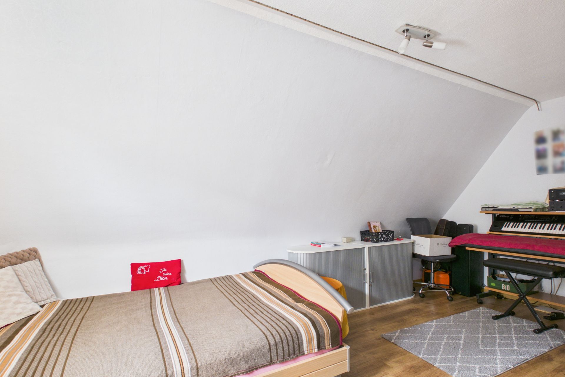 Zimmer mit Einzelbett, Teppich, Schreibtisch und E-Piano unter einer Dachschräge.