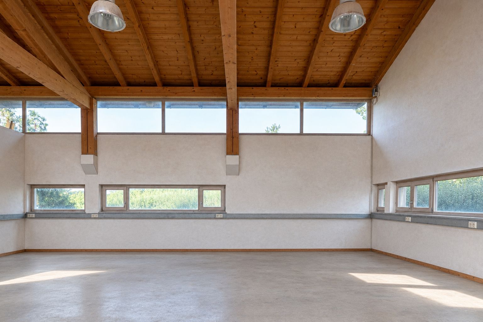 Leerer Raum mit hohen Decken, Holzbalken und vielen Fenstern an den Wänden.