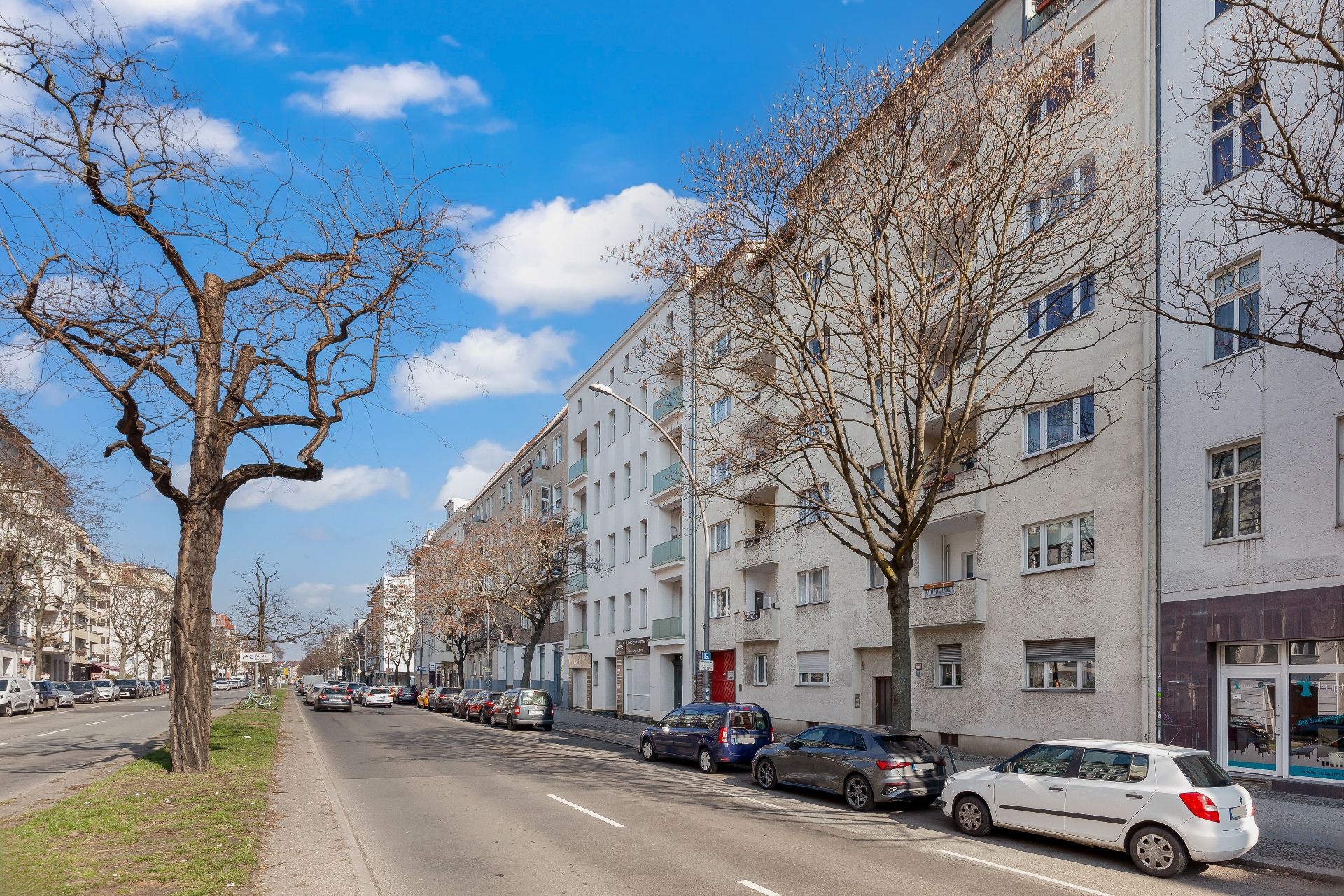 Mehrstöckiges Wohnhaus an einer Straße mit geparkten Autos und kahlen Bäumen.