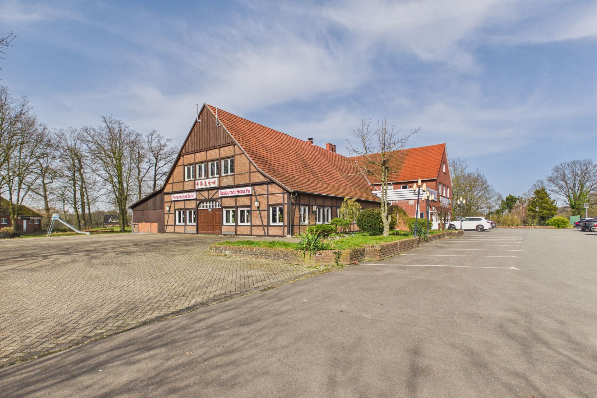Großes Fachwerkhaus mit rotem Dach und angrenzendem Parkplatz, Bäume im Hintergrund.