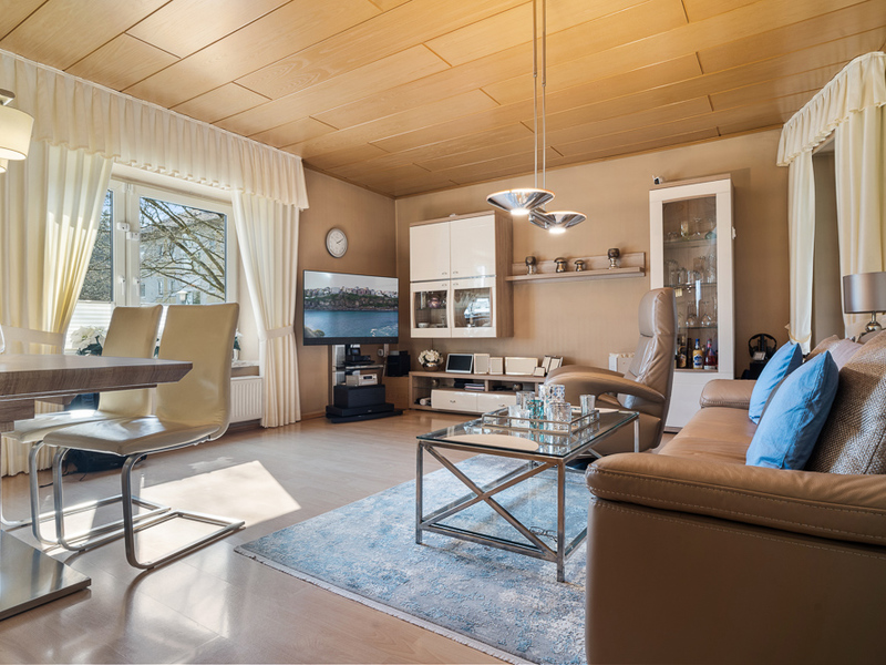Helles Wohnzimmer mit Ledersofa, Glastisch, TV, Schrankwand und großem Fenster mit Vorhängen.
