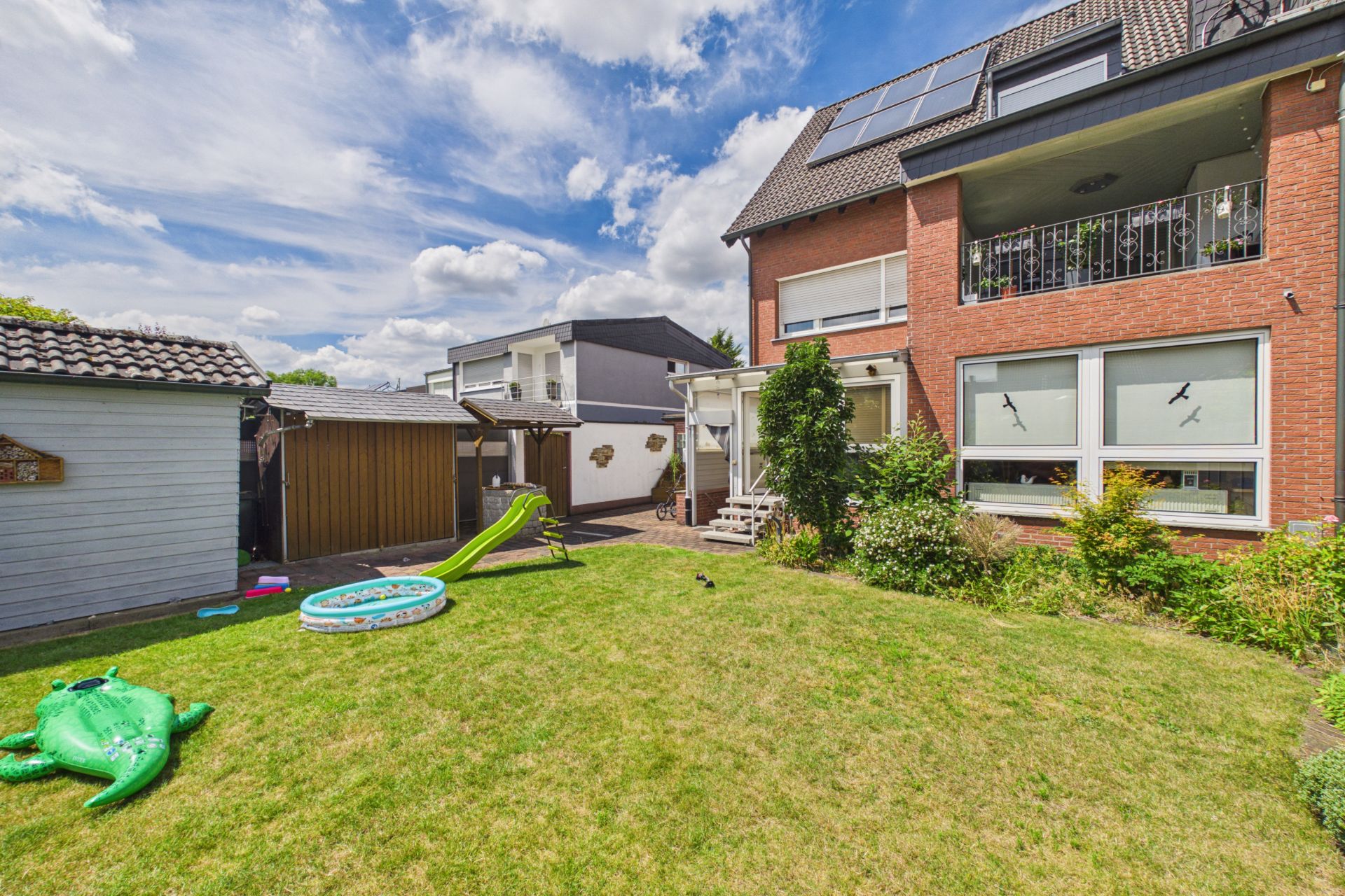 Garten mit Rasen, aufblasbarem Pool, Rutsche und grünem Spielzeug, angrenzend an ein Wohnhaus.