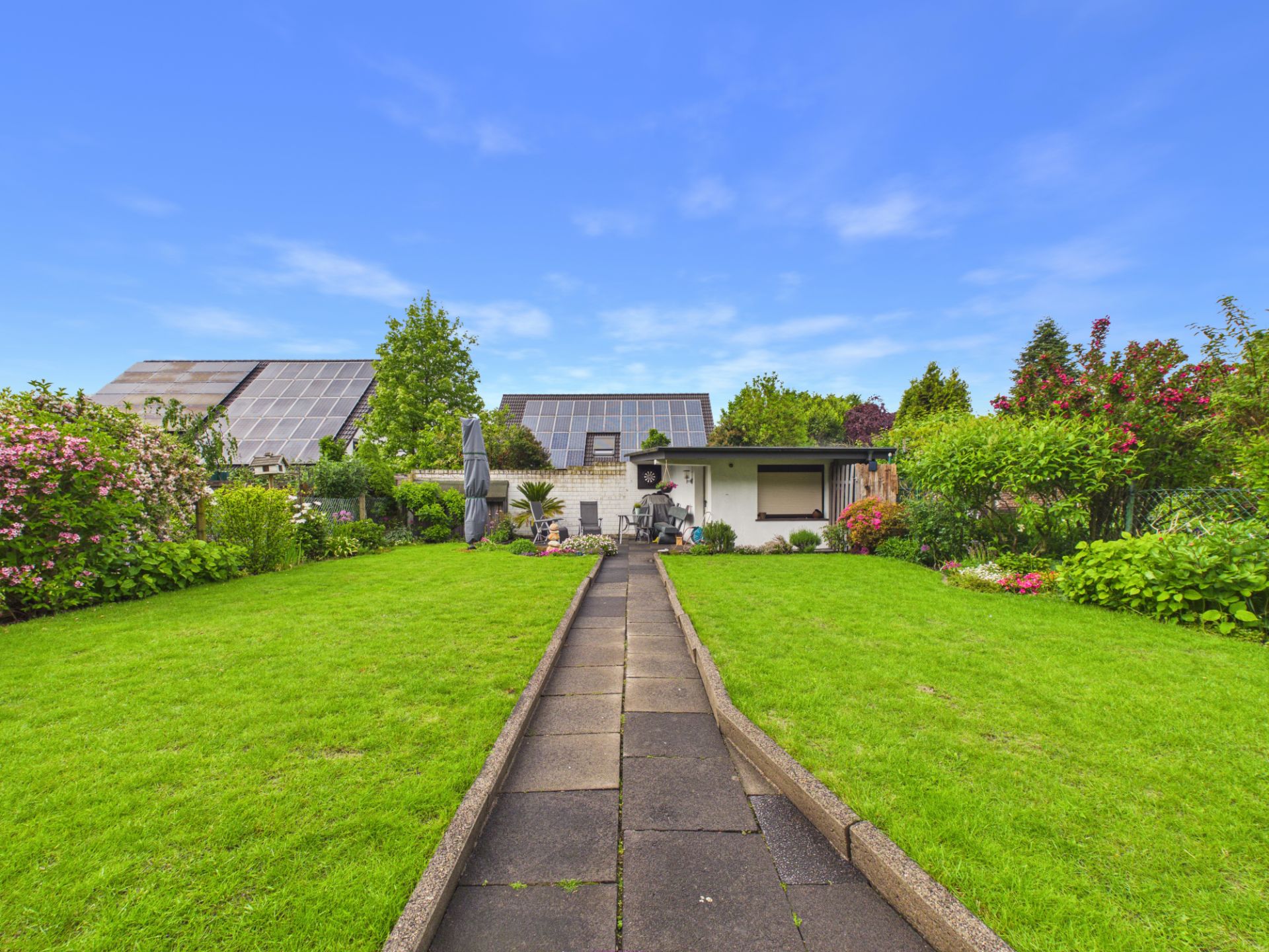 Gepflegter Garten mit Rasen, Blumenbeeten und Weg zu einem Haus mit Terrasse.