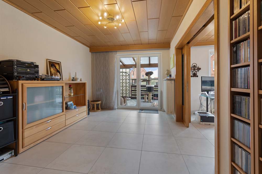Wohnzimmer mit Holzmöbeln, Fliesenboden, Deckenleuchte und Zugang zur Terrasse.