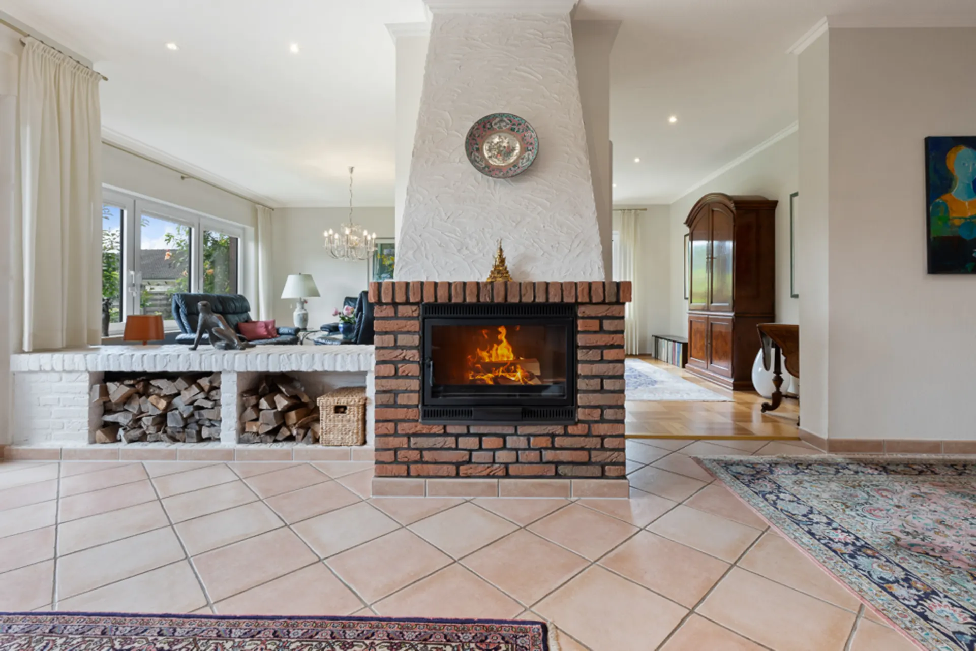 Wohnzimmer mit Kamin, Holzlager, großen Fenstern und Teppichen auf Fliesenboden.