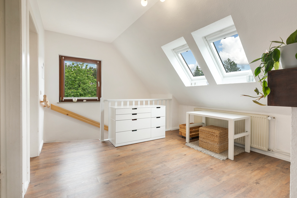 Heller Raum mit schrägen Fenstern, Kommode, Tisch, Pflanzen und Holzfußboden.