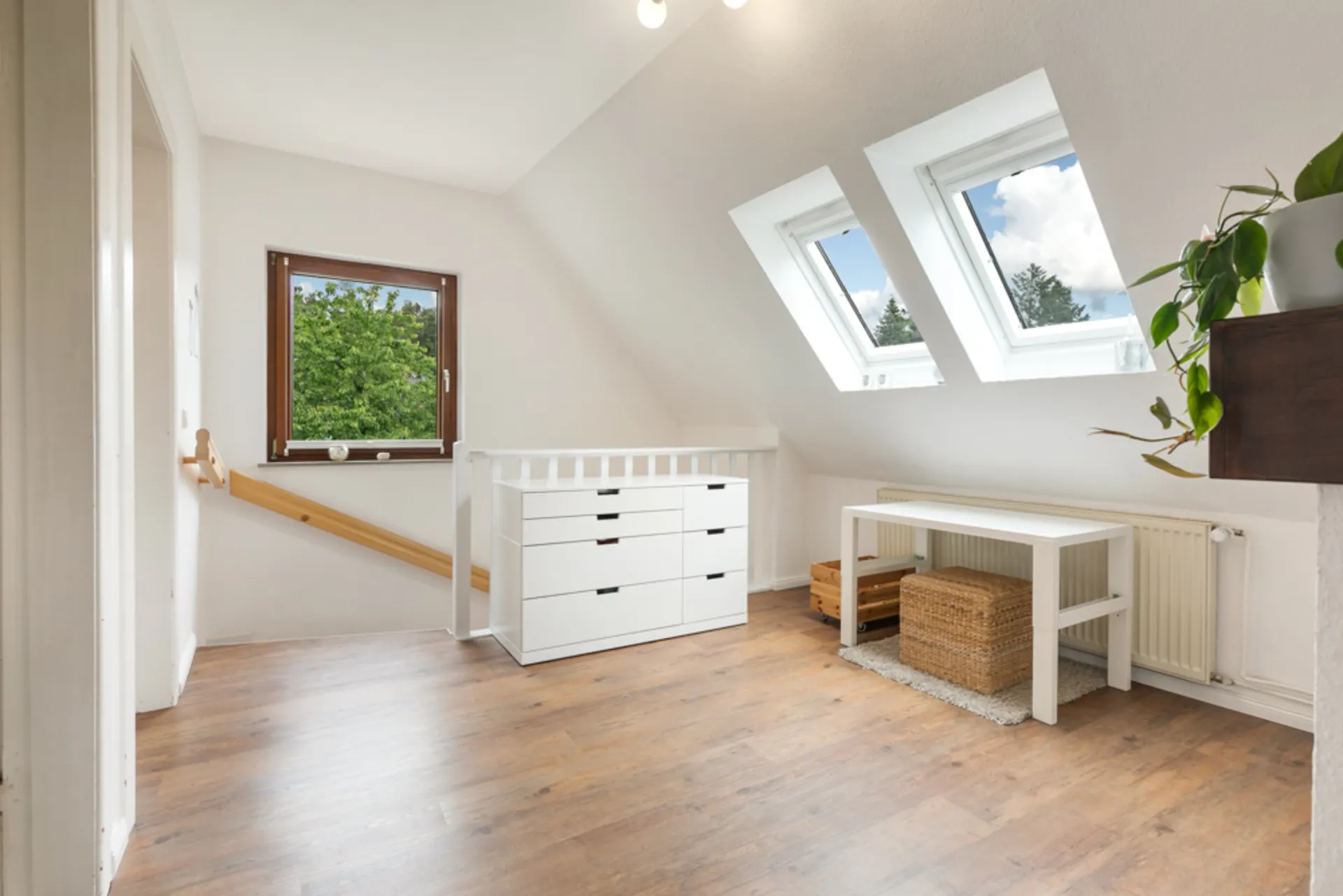 Heller Raum mit schrägen Fenstern, Kommode, Tisch, Pflanzen und Holzfußboden.