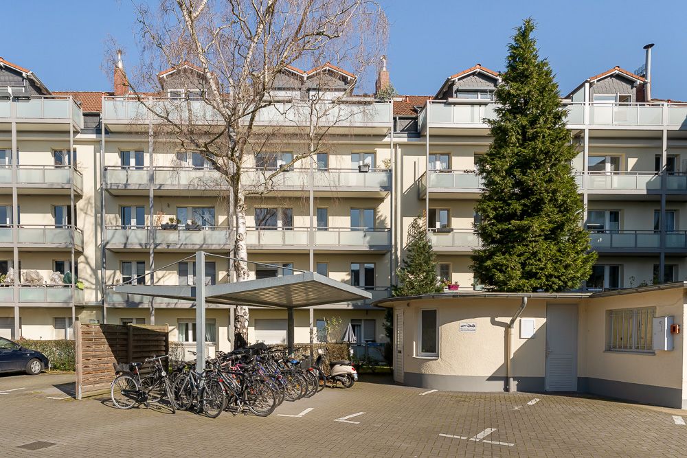 Mehrstöckiges Wohnhaus mit Balkonen, davor Fahrradständer und große Bäume.